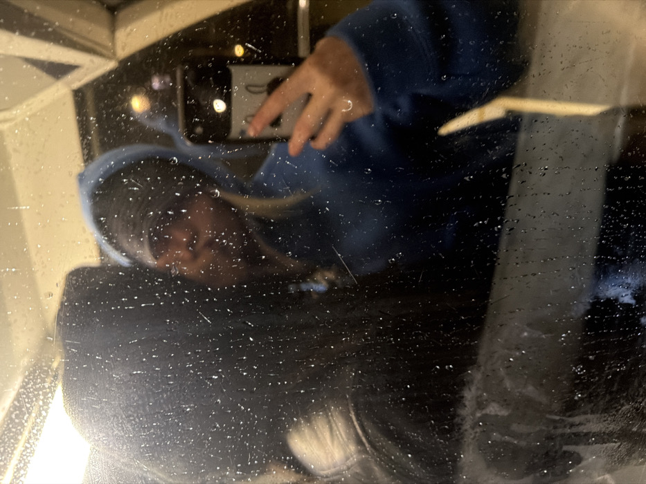 A blurry, distorted reflection shows a person wearing a blue hoodie, seemingly taking a photo with a phone. The reflection is obscured by water droplets on a glass or reflective surface. A hand holding the phone is visible in the upper portion of the reflection. The lighting is dim and the overall image has a grainy, low-resolution quality.