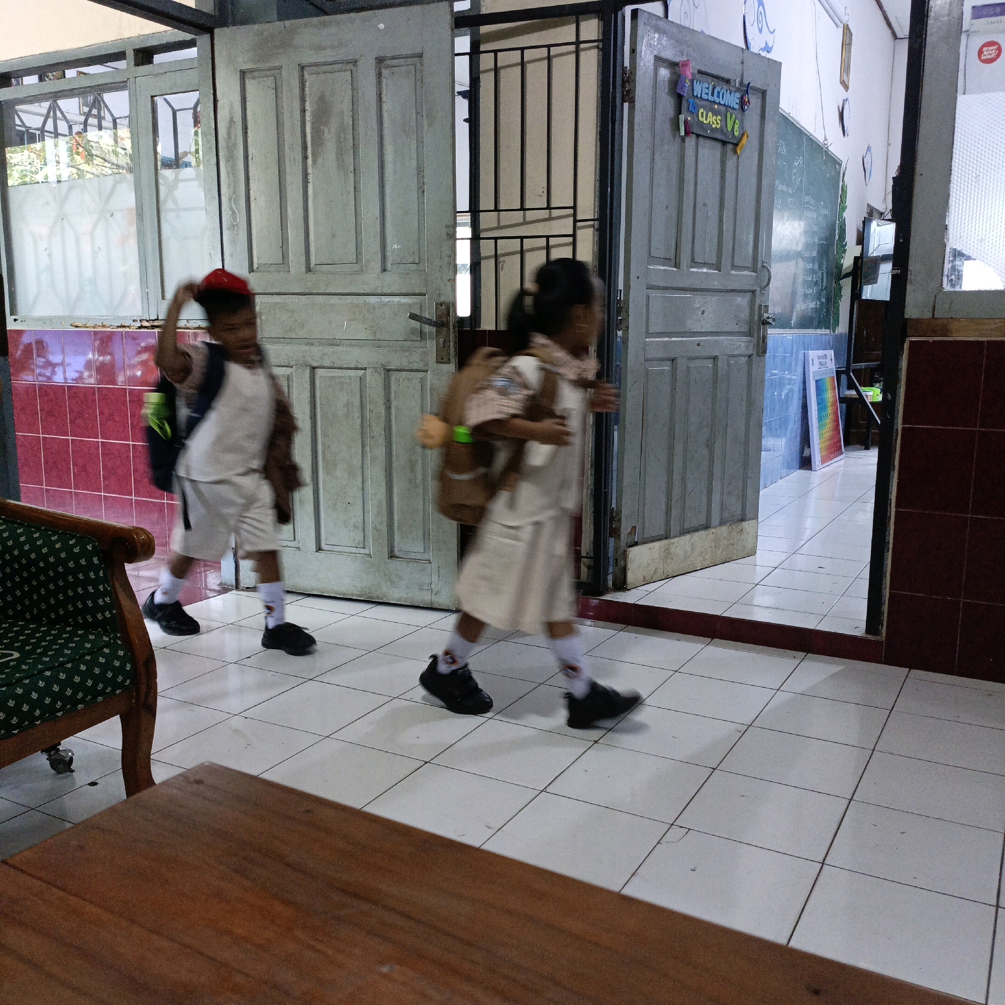 Two children, a boy and a girl, are walking in a tiled hallway inside a building in Salatiga, Indonesia. The boy, on the left, wears a red cap, a white shirt, and white shorts, carrying a backpack. The girl, on the right, wears a light-colored uniform and also carries a backpack. They are both blurred due to motion. The hallway has grey double doors on the left and a slightly ajar grey door on the right with a "WELCOME TO CLASS V8" sign. The walls are tiled in red and white, and a blackboard is visible through the open doorway on the right. A wooden table and a green armchair are partially visible in the foreground.
