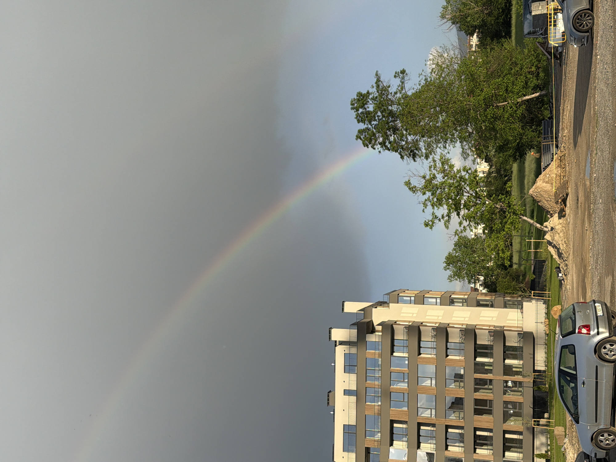 Here is a description of the image:
The image shows a residential area with a rainbow in the sky. A modern apartment building with large windows is in the foreground. A silver car is parked near the building. In the background there is greenery, including trees and grass. The sky is mostly overcast, with a distinct rainbow arcing across it. The location appears to be a city, given the presence of the apartment building and the car. The city and state are not identifiable from the image.