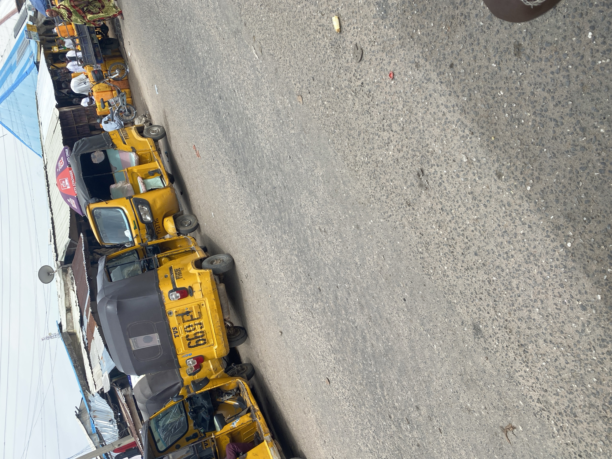 The image depicts a street scene in Jalingo, Nigeria, characterized by a row of yellow motorized rickshaws, also known as *keke napeps*. The rickshaws are parked along the side of an asphalt road, which appears well-trafficked with a rough texture. The scene suggests a bustling environment, likely a commercial area or a transport hub.
Several individuals are visible in the background, some tending to stalls or engaging in other activities. One person is seated in the driver's seat of one of the rickshaws. The overall atmosphere seems active and typical of a daytime scene in a Nigerian city. The weather appears to be fair, with no indication of rain or extreme conditions.
Notable details include the vibrant yellow color of the rickshaws, which are a common mode of transport in the region. Some of the rickshaws have covers over the passenger areas. There is also a blue and white striped awning visible on the left side of the image, possibly belonging to a shop or stall.
Visible text includes the brand name "TVS" on one of the rickshaws, along with license plates and other markings that are difficult to read due to the image's orientation and resolution. One rickshaw has "GETS STRONGER" written on its canopy. The license plate on one of the vehicles clearly shows "PT-377G". Another visible license plate is "6699 FGN".
The image is oriented vertically, with the ground and road occupying the majority of the frame. The perspective is from a slightly elevated viewpoint looking down at the parked vehicles and the street.