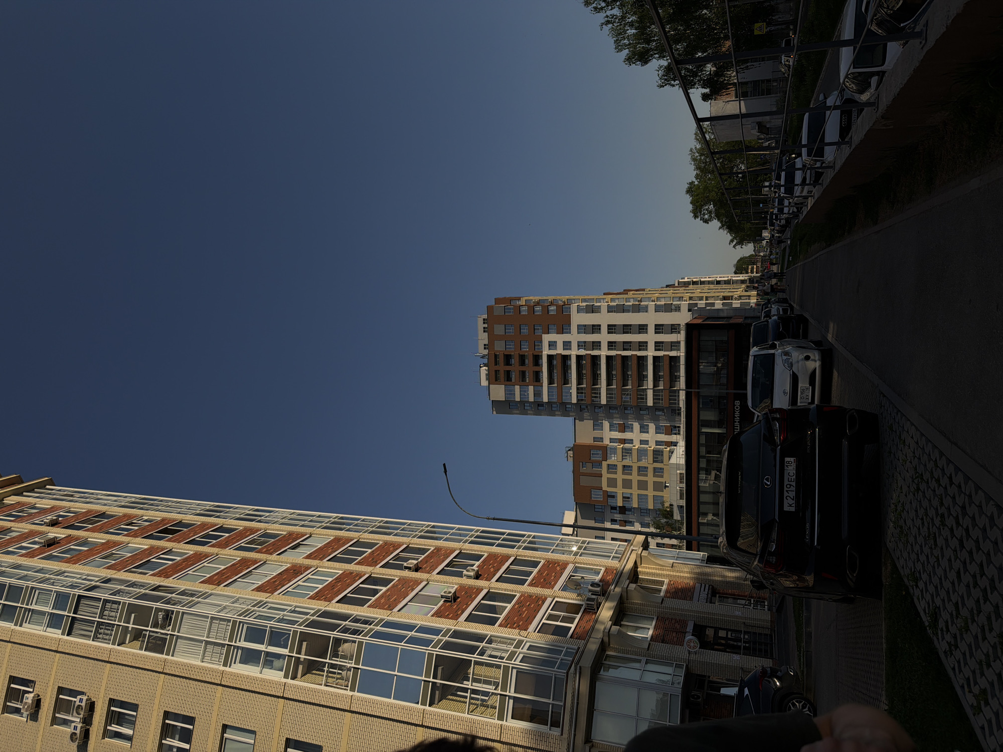 The image depicts a street in Izhevsk, Russia. On the left side of the road, there is a multistory building made of light-colored brick with rows of windows separated by red brick accents. To the right and farther into the distance, there is a taller high-rise apartment building that is primarily white and brown.
Parked along the right side of the street is a line of cars. A black Lexus is parked closest to the viewer with a license plate reading "K219EC 18." Beyond that, several other cars of varying makes and models are parked alongside the curb. The right side of the road also features a sidewalk bordering the street.
The sky is clear and blue. A tall lamppost is visible near the base of the first building. Trees are visible at the top-right corner, with some greenery present next to the sidewalk.