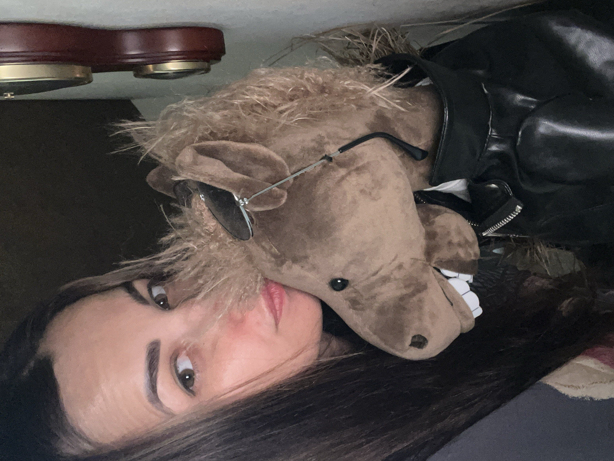 A medium shot captures a woman with long, dark brown hair and brown eyes, looking at the camera from the left side of the frame. She is holding a plush toy horse, which is brown with a tan mane and wearing aviator sunglasses and a black leather jacket. The horse's mouth is open, revealing white teeth. The background is dark and out of focus, with a portion of a wall and two decorative clocks visible on the right side of the frame. The lighting is dim, creating a close-up, intimate feel.