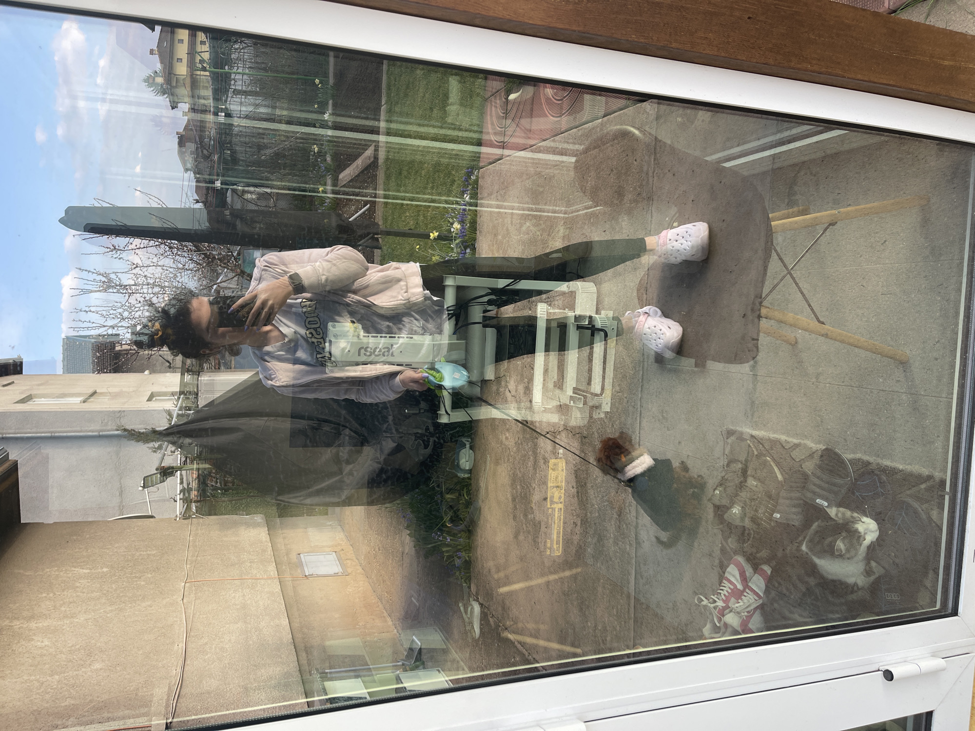 The image depicts a view through a glass window, reflecting both an interior and exterior scene. Inside, a woman is seen holding a phone, possibly taking a picture of herself or the reflection. Outside, there's a yard with a small dog on a leash, some greenery, and a chair. The reflection shows buildings typical of an urban environment, suggesting a city. It is difficult to identify the location as Sofia, Bulgaria without further information. However, based on the style of buildings and urban landscape, it is probable that the location is Sofia.