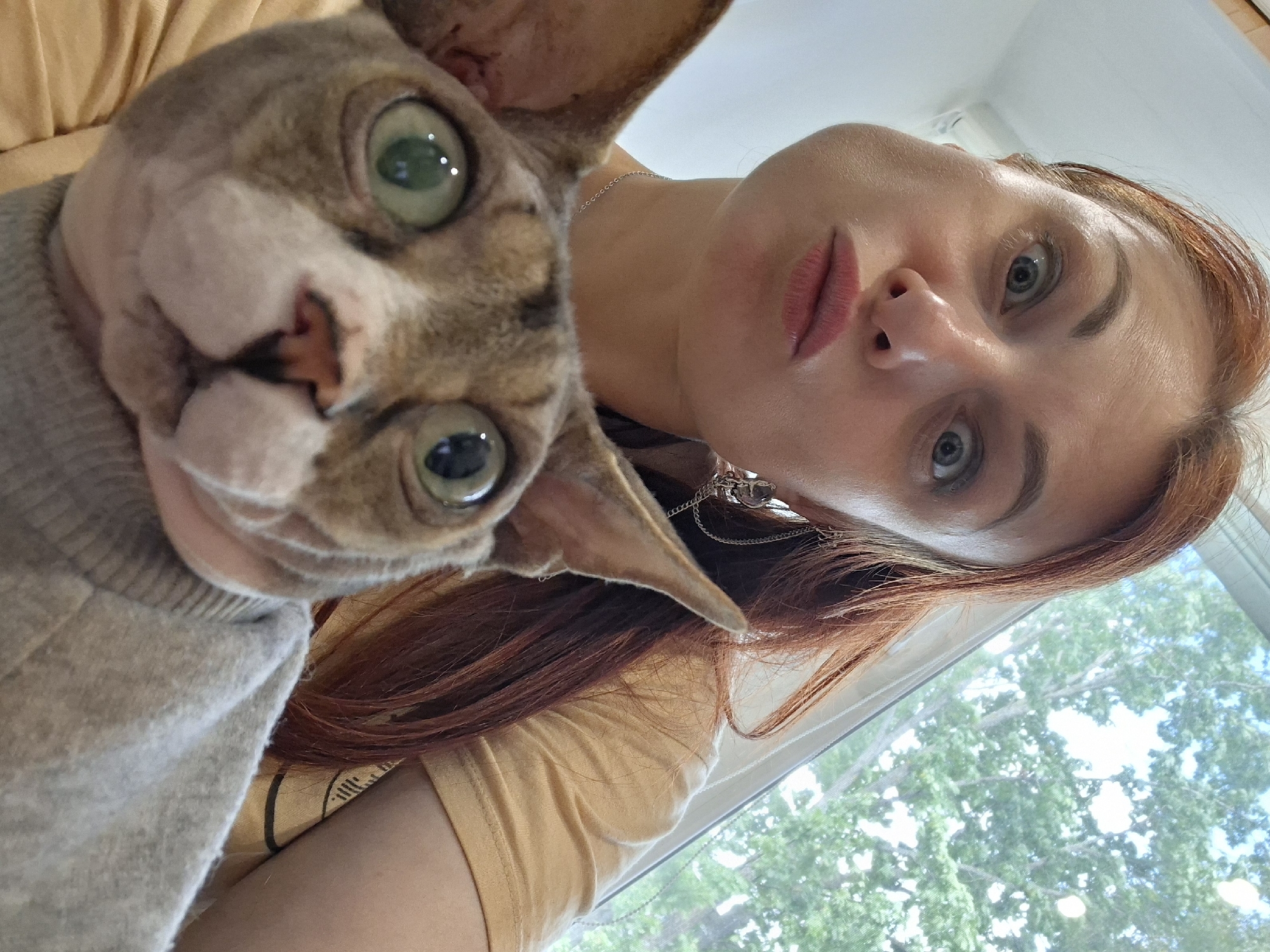 A close-up, low-angle selfie shows a woman with red hair and a surprised expression taking a picture with a Sphynx cat. The woman is looking at the camera with wide eyes and her lips pursed. The cat, also looking at the camera, has large green eyes and is wearing a grey sweater. The background is blurred, showing a window with trees and greenery outside. The woman is wearing a yellow t-shirt and a silver chain necklace. The image appears to be taken indoors, likely in a residential setting in Myadzyel, Belarus.