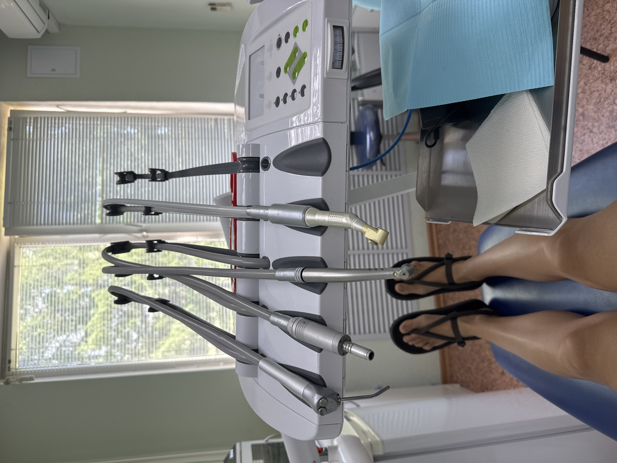 The image shows a dental operatory. A dental unit with various handpieces and controls is prominently displayed. A patient's legs are visible in the lower right, clad in black sandals and seated in the dental chair. The room is illuminated by natural light filtering through a window in the background.