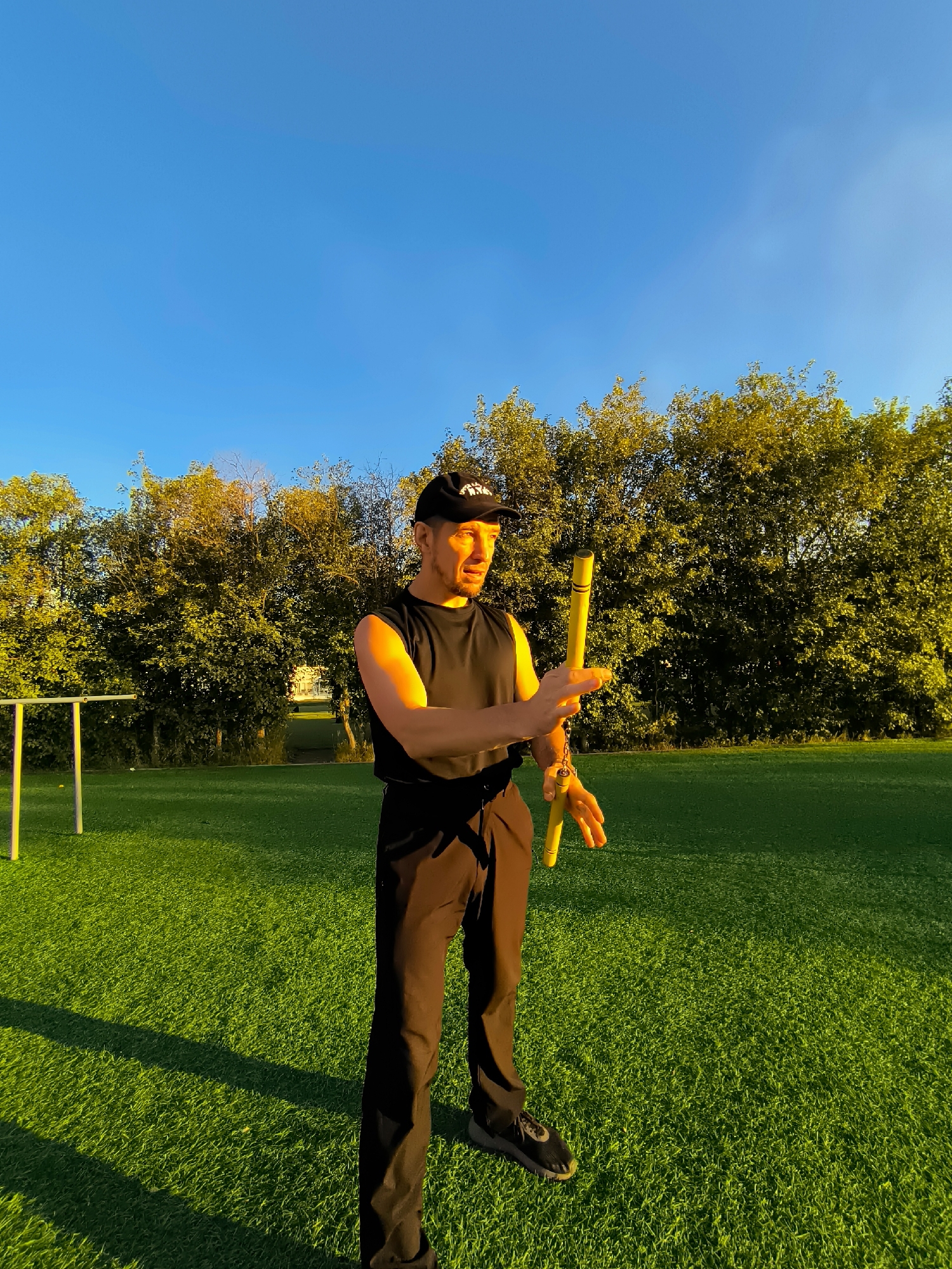 A man wearing a black tank top, black baseball cap, and black pants stands on a bright green turf field, holding a yellow nunchaku. He is facing left, with his right arm bent and his left hand extended. The background features a dense line of trees with sun-drenched leaves, set against a clear blue sky. To the left of the man, a portion of a white and yellow sports apparatus is visible. The lighting suggests it is late afternoon or early evening, with long shadows cast across the field. The location is identified as the city of Qarağandı, Kazakhstan.