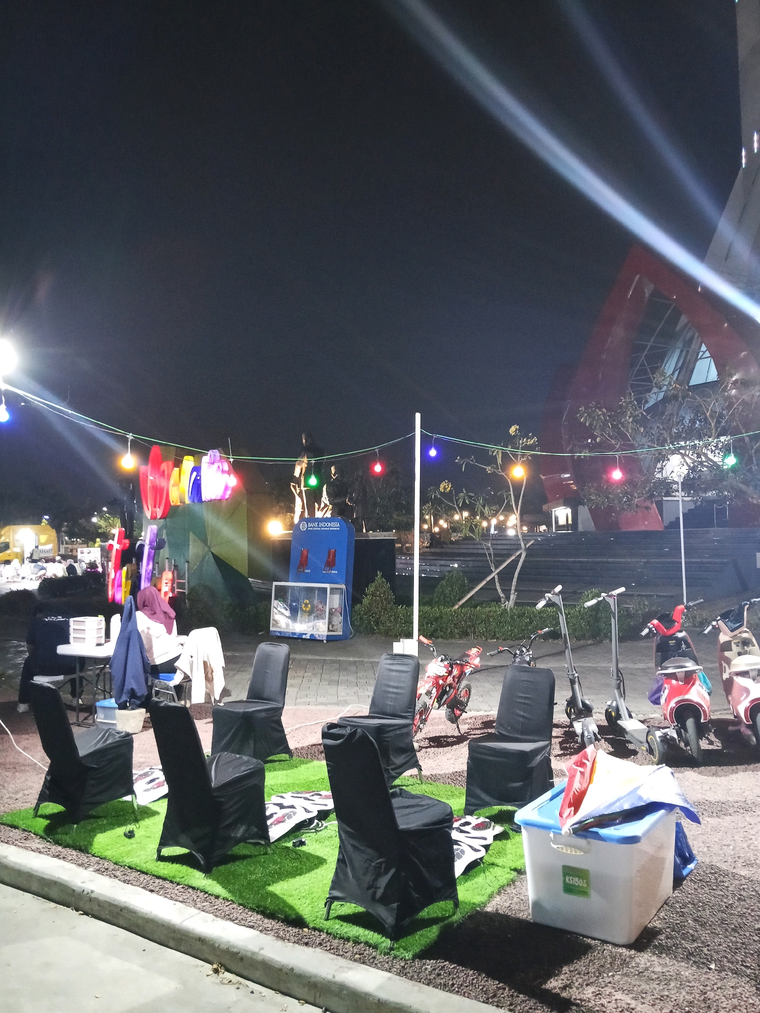 The image depicts an outdoor night scene in the city of Purwokerto, Indonesia. In the foreground, several chairs covered in black fabric are arranged on a patch of artificial grass. To the right, a white plastic bin with a blue lid is visible, containing colorful items. Behind the chairs, there are several electric scooters and mopeds parked. In the background, a large building with a distinctive red architectural feature is illuminated. Strings of colorful lights are strung across the area, and spotlights cast beams into the night sky. A blue booth labeled "BANK INDONESIA" is also present, along with a table and chairs where two individuals are seated. The overall atmosphere suggests an event or gathering taking place outdoors at night.
