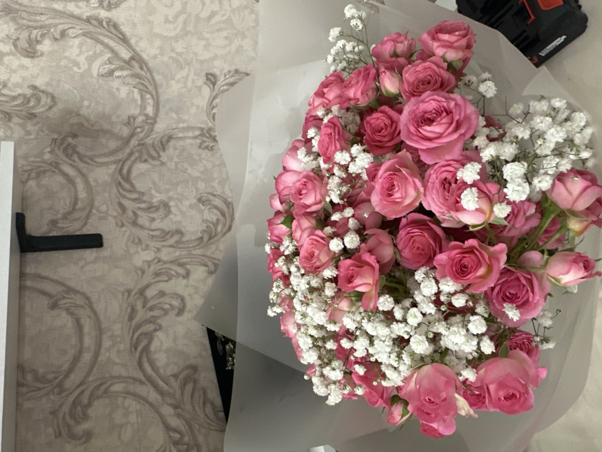 A bouquet of pink roses and white baby's breath is presented in a white paper wrap. The bouquet is arranged in front of a wall with a subtle, ornate wallpaper pattern featuring scrolling designs in a neutral color palette. A black bracket is visible on the wall above the bouquet. The lower right corner of the image shows a portion of a black object with an orange accent, possibly electronic equipment. The scene appears to be indoors, likely a room in a home or establishment. There are no people present in the image.