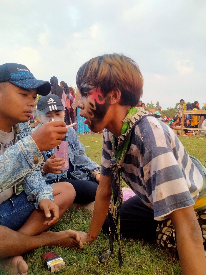 The image depicts an outdoor gathering on a grassy field in the city of Glonggong, Indonesia, under an overcast sky. In the foreground, three individuals are seated on the grass. To the left, a barefoot male wearing a blue and black baseball cap and a denim jacket extends his left hand, holding a white cigarette. A female next to him, wearing a black Adidas cap and a denim jacket, lights this cigarette with a lighter held in her left hand, producing visible smoke. Her right hand holds a pink rectangular object. An open pack of Marlboro cigarettes rests on the grass nearby. To the right, a third male, also barefoot, is seated, wearing a white and blue striped t-shirt with visible stains, a patterned neck scarf, and face paint featuring black and red patterns around his left eye. He is looking towards the other two individuals with an open mouth. Numerous other people are present in the blurred background, some standing and some seated, with a wooden structure visible to the far right, indicating a public event.