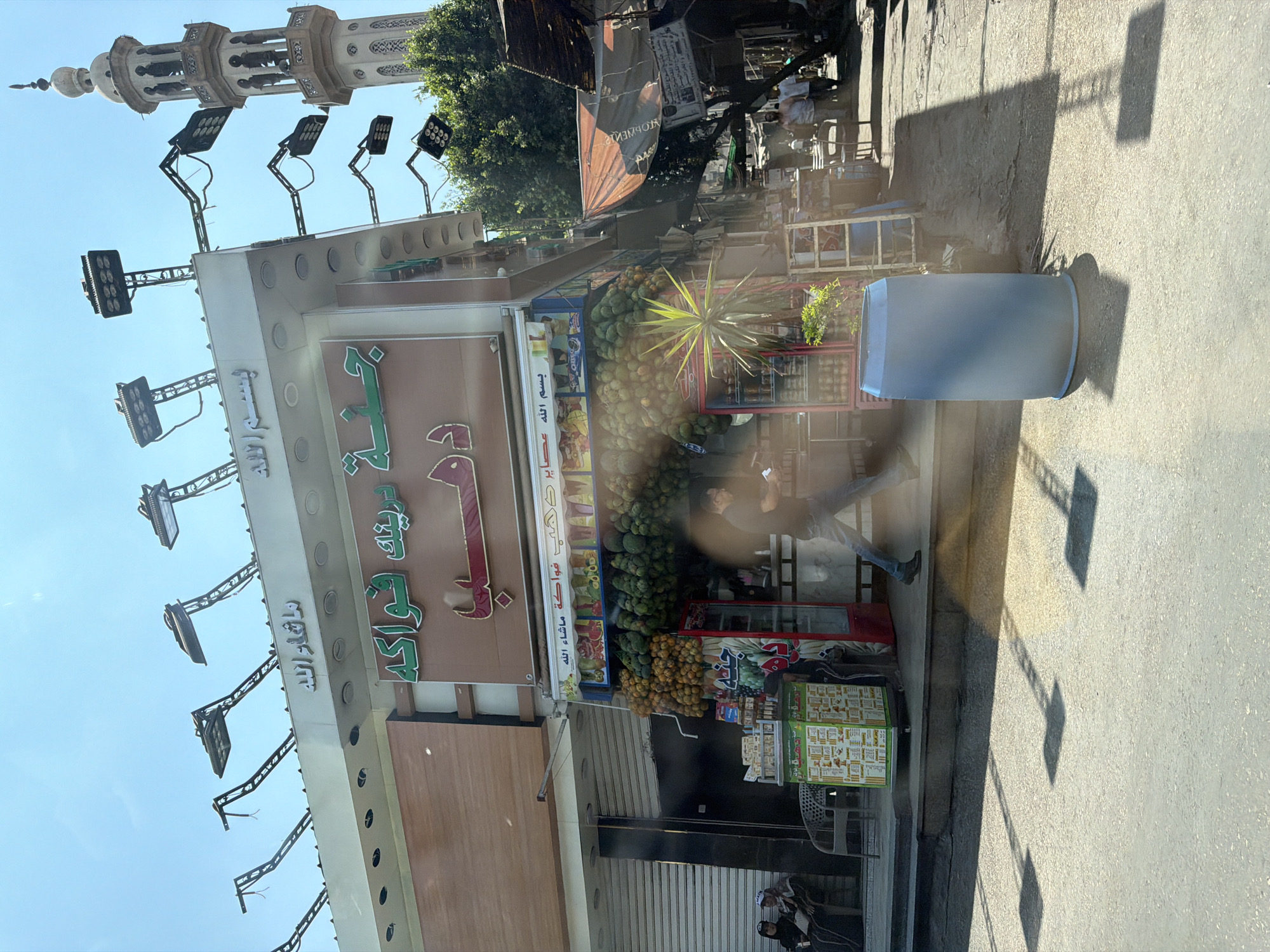 The image captures a sunlit street scene, likely in Cairo, Egypt, with a juice and fruit shop as the central focus. The shop's facade is adorned with Arabic signage, including "بسم الله" (In the name of God) and "ما شاء الله" (What God has willed). Above the shop, a row of modern, industrial-style lights with multiple LED panels are mounted on angled metal arms extending from the roof, some with dangling wires. To the right, a traditional minaret of a mosque rises into the clear blue sky.
The shop itself displays an abundance of fresh produce, including large quantities of green avocados and smaller, orange fruits resembling tangerines or small oranges, meticulously arranged. A red-fronted refrigerator is visible behind the fruit, stocked with beverages. A green display stand with menus and product information is positioned to the left of the entrance.
In the foreground, a person in dark clothing is captured in motion, walking across the street, their legs blurred by the movement. The pavement is marked with distinct square shadows, likely cast by the overhead lights. A large, light blue cylindrical bin stands prominently on the right side of the street. In the background, partially obscured by trees and awnings, other shops and individuals are visible, suggesting a busy marketplace. Two women in traditional Islamic attire are seated on a bench to the left of the juice shop, observing the street. The overall impression is one of vibrant street life within an urban Egyptian setting.
