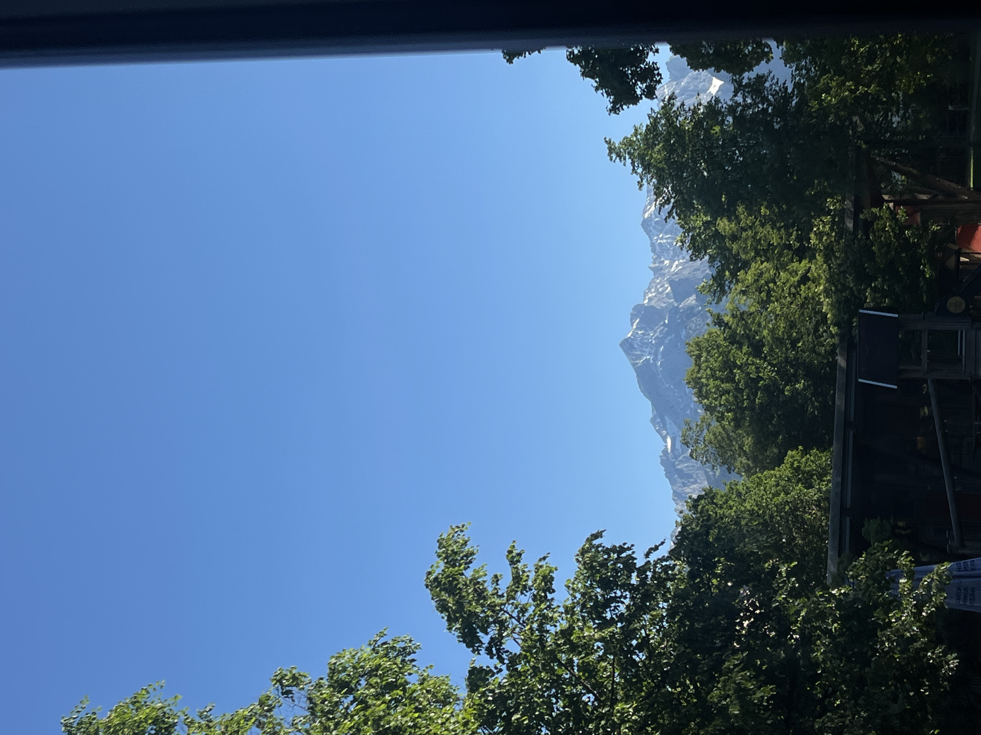 The image shows a view of the sky and mountains framed by trees. The sky is a clear blue, taking up the majority of the frame. Trees with green leaves occupy the lower and right portions of the image. In the distance, a snow-capped mountain range is visible. The photograph appears to be taken from an elevated position, with part of a frame or window visible along the top edge. The location is likely Garmisch-Partenkirchen, Germany, given the presence of the Bavarian Alps, although specific details connecting it directly to Garmisch-Partenkirchen are absent. No people or interactions are visible.