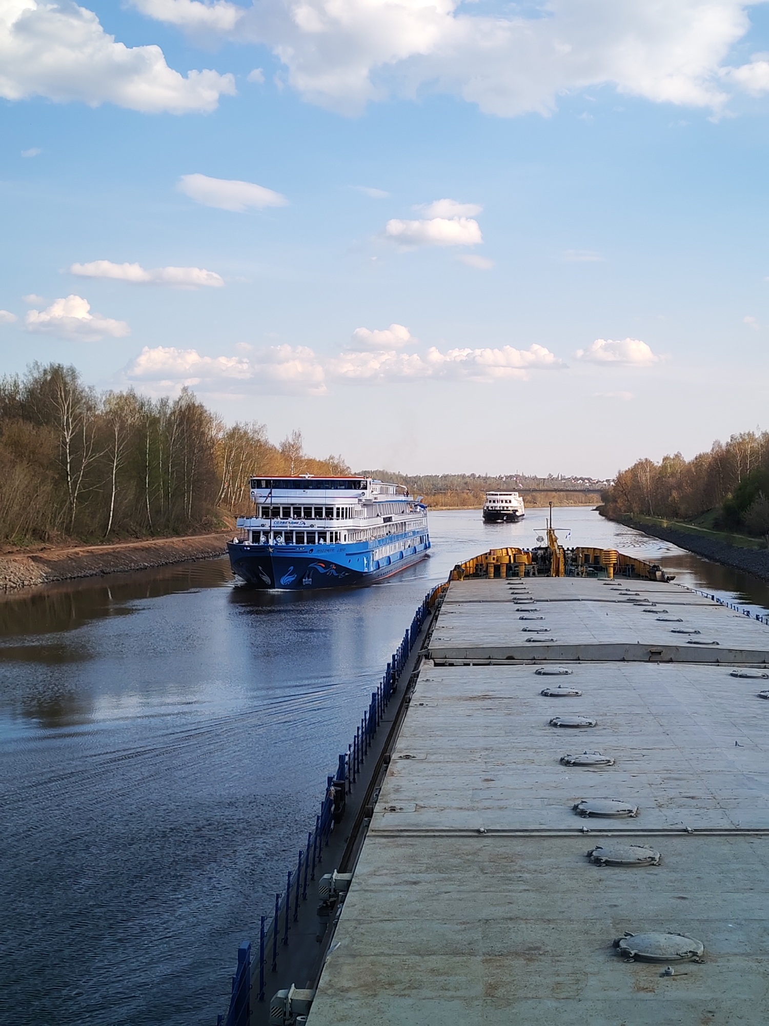 The image shows a canal with two cruise ships passing. The viewer is positioned on a large barge or freighter. The sky is light blue with puffy white clouds. Trees line the banks of the canal.