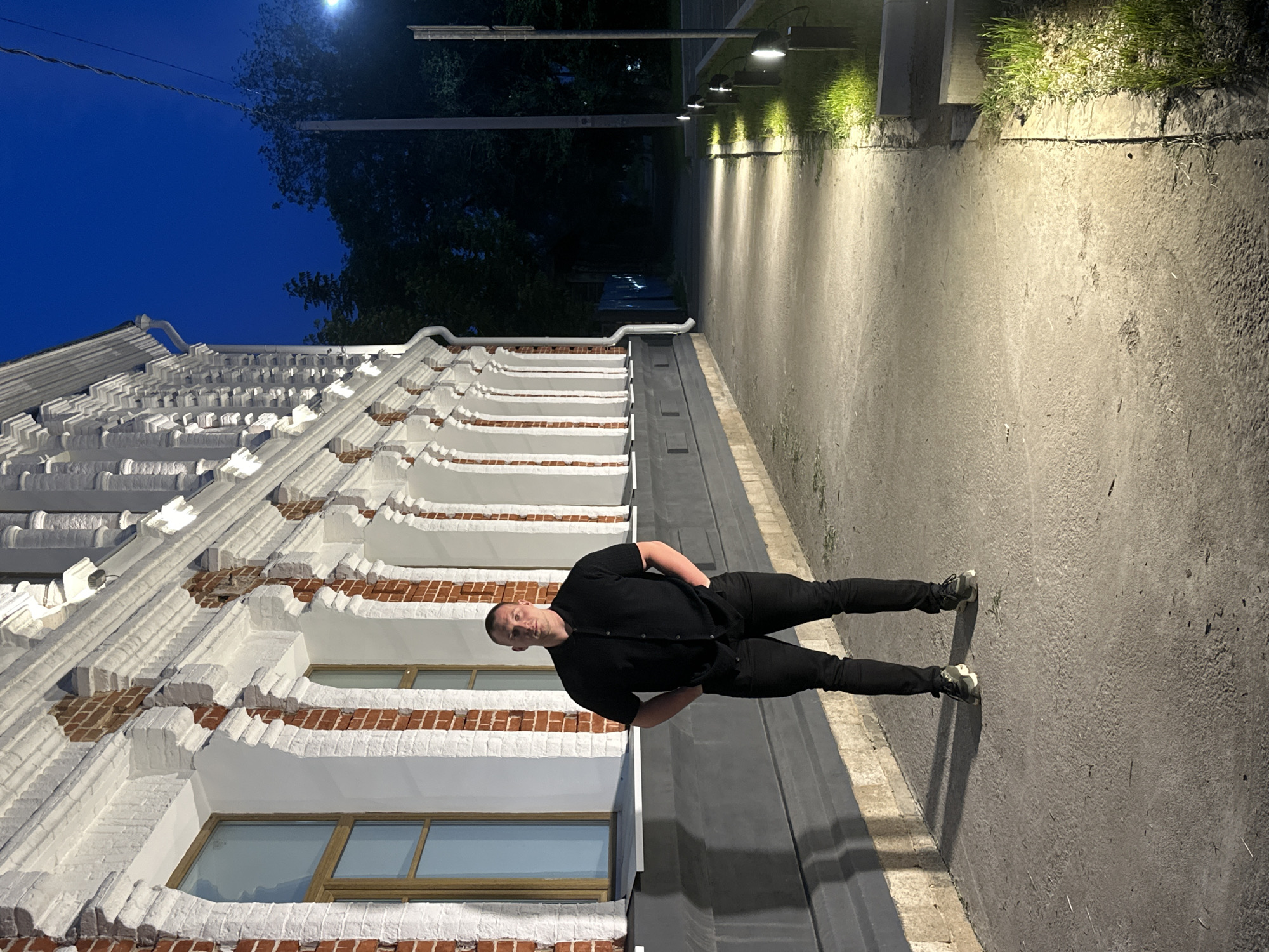 A full-body, eye-level shot captures a man standing on a paved pathway next to a building at night. The man is centered in the frame, facing forward with his hands in his pockets. He is wearing a black collared shirt and black pants, along with dark sneakers. The building to his left features red brick and white decorative elements, with several arched windows. The pathway is illuminated by several low-lying lights, casting a warm glow and creating shadows. To the right of the pathway, there is a strip of grass and more lights. Beyond the pathway and grass, trees are visible in the background, silhouetted against the dark blue night sky. The scene suggests an urban or semi-urban environment, possibly in Blagoveshchensk, Russia, given the context of the request.