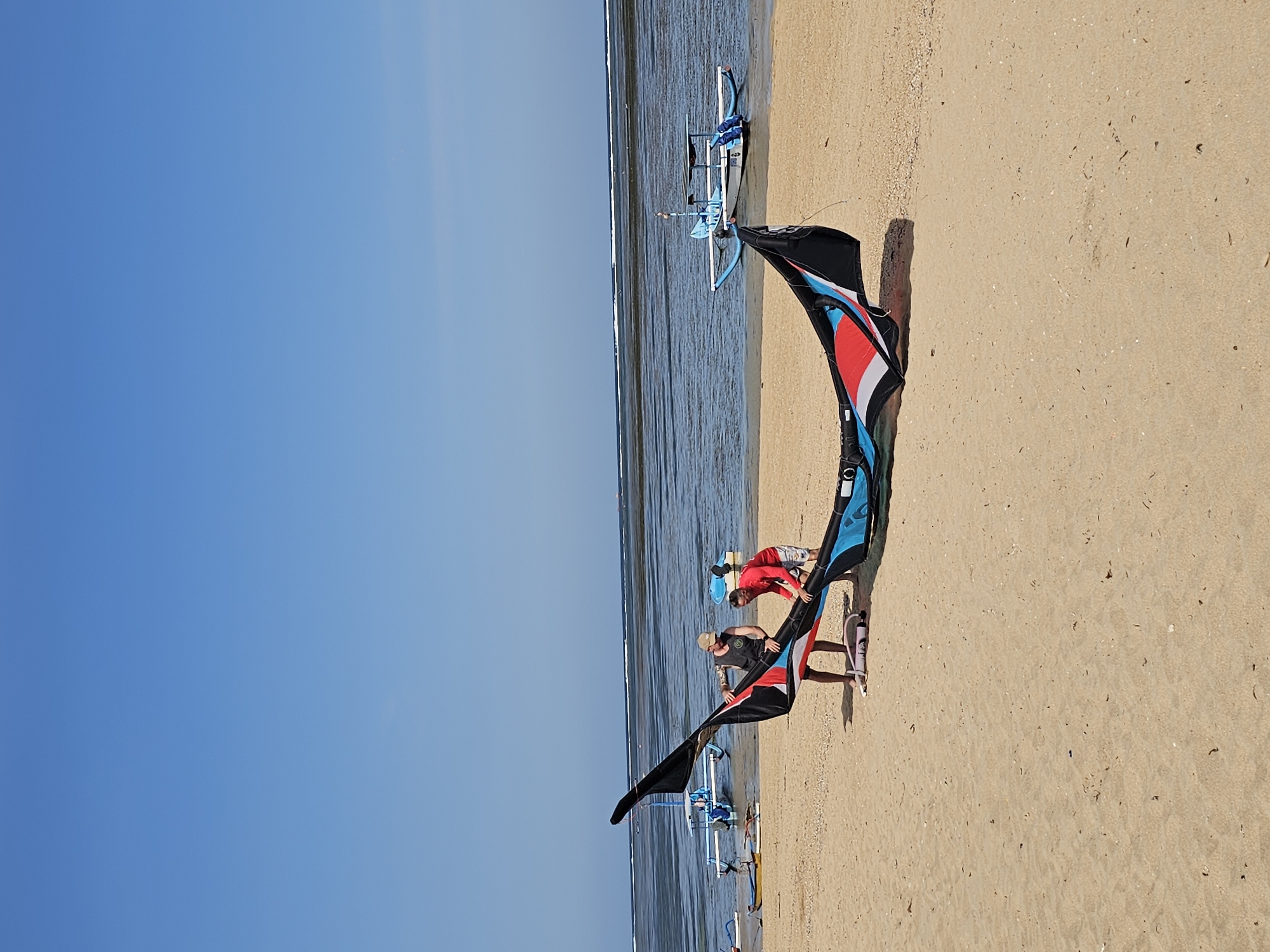 This image, taken at Padang Galak Beach in Denpasar, Indonesia, depicts a bright, sunny day with a clear blue sky. Two individuals are standing on a sandy beach, working together to assemble a large, colorful kite. The kite, predominantly black with accents of red, white, and blue, is partially unfurled on the sand. One person, wearing a black tank top and a beige cap, is on the left, and the other, in a red t-shirt and patterned shorts, is on the right. Both appear to be engaged in the process of setting up the kite.
In the background, the ocean stretches out, appearing calm with a few small waves breaking closer to the shore. Several traditional Indonesian outrigger boats, known as *prahu*, are visible in the shallow water. Some are moored near the beach, while others are further out. One prominent blue and white *prahu* with a canopy is closest to the shore. The overall scene suggests preparations for water sports, likely kitesurfing, given the kite's presence. The sand is light brown and appears to be a mix of fine grains and small shell fragments. The tide seems to be low, exposing a wide expanse of wet sand and shallow water.