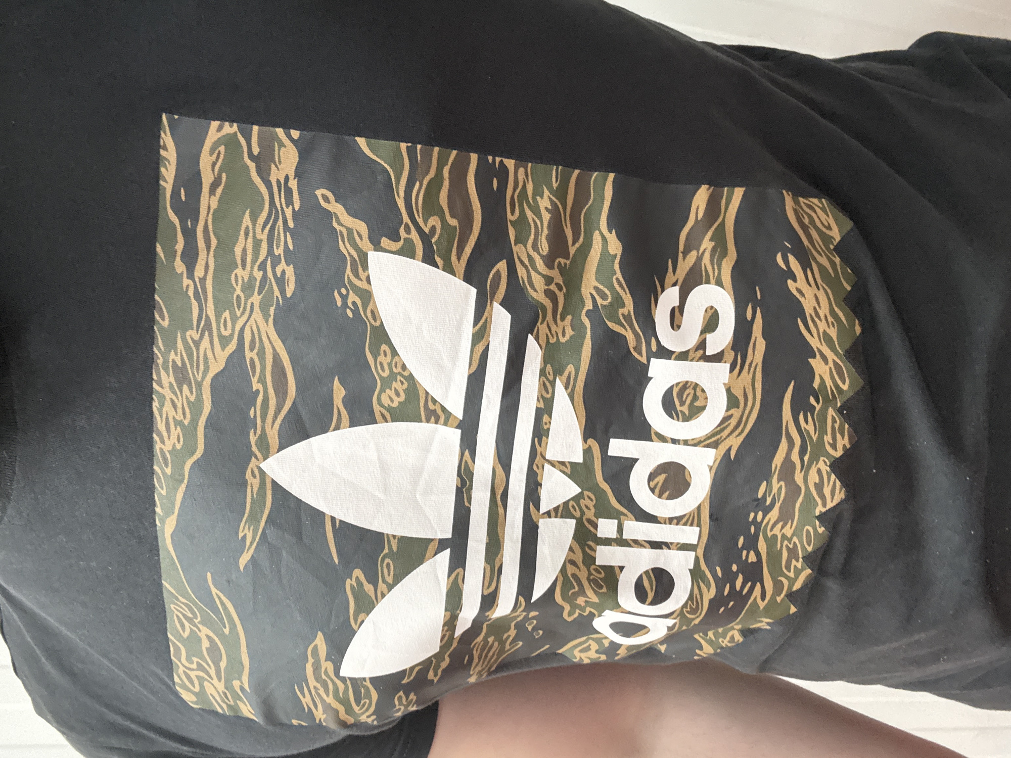 The image presents a close-up view of a person wearing a black Adidas t-shirt, presumably indoors in Stara Zagora, Bulgaria. The shirt features a prominent graphic that incorporates a distinctive camouflage pattern as its background. This pattern is composed of intricate, wavy, and abstract shapes in shades of dark green, dark grey, and a lighter brown or khaki, giving it a "tiger stripe" aesthetic.
Overlaid on this camouflage pattern are the white Adidas trefoil logo and the "adidas" wordmark. The trefoil logo, characterized by its three leaf-like sections formed from parallel stripes, is positioned on the left side of the graphic. To its right, the "adidas" text is displayed vertically, rotated 90 degrees clockwise, with each letter clearly visible against the patterned backdrop.
The black fabric of the t-shirt is visible around the edges of the central graphic and exhibits some minor creasing, indicating it is being worn. The bottom of the frame shows a small portion of the wearer's skin. The background is a plain, light color, suggesting an indoor setting. The image is well-lit, providing clear details of the t-shirt's design and texture, with a strong focus on the brand's logo and the unique camouflage print.