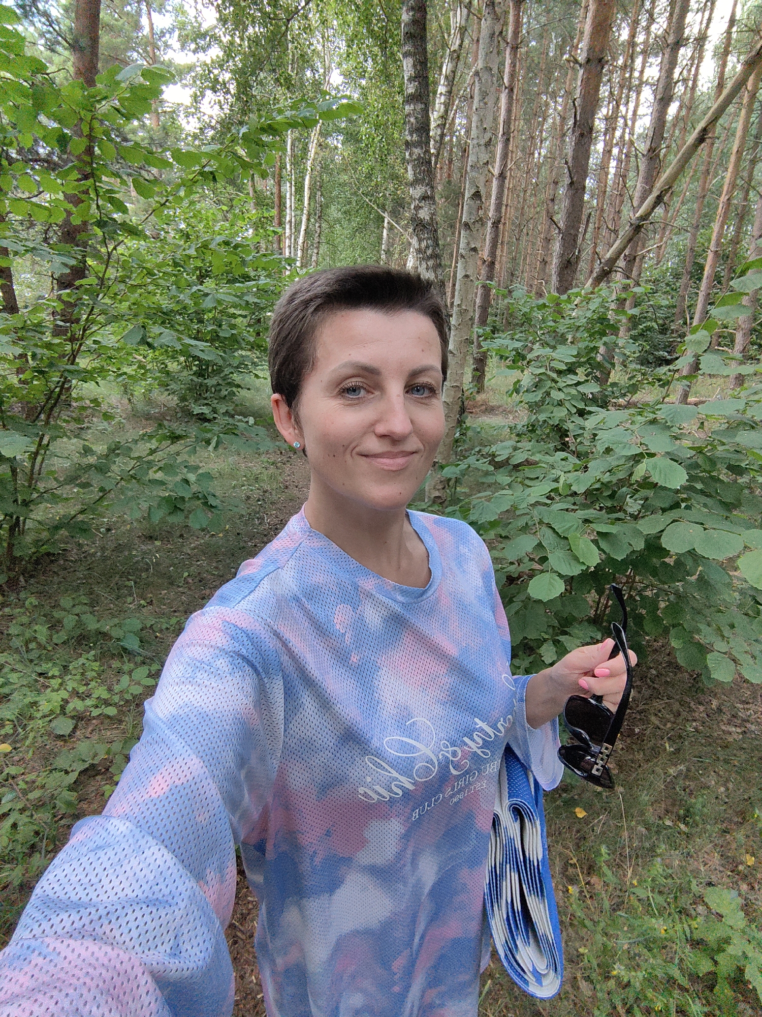 A young woman with short, dark hair and blue eyes takes a selfie outdoors. She is wearing a light blue, pink, and white tie-dye long-sleeved shirt made of mesh fabric. Her left arm is extended to hold the camera, and her right hand is holding a pair of black sunglasses and a rolled-up blue and white item. She is smiling at the camera. The background is a forest with tall trees and dense green foliage. The ground is covered with grass and dry leaves. The scene appears to be in a park or wooded area, possibly in the Мядзельскі сельскі Савет region of Belarus, given the types of trees and vegetation.