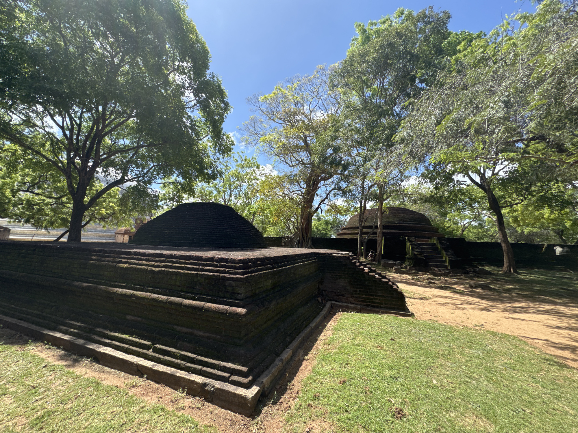 The image depicts a brick structure, likely an ancient ruin, situated in a grassy area with trees, probably a park or historical site. The ruin consists of a tiered, rectangular base topped with a dome-shaped structure. In the background, another similar brick structure with steps is visible, also dome-shaped, surrounded by trees. The setting seems to be in an outdoor environment with a clear sky. The scene strongly suggests a location in the ancient city of Polonnaruwa, Sri Lanka, given the architectural style of the brick structures, which are reminiscent of the city's well-preserved ruins. There is no presence of people or action in the scene.