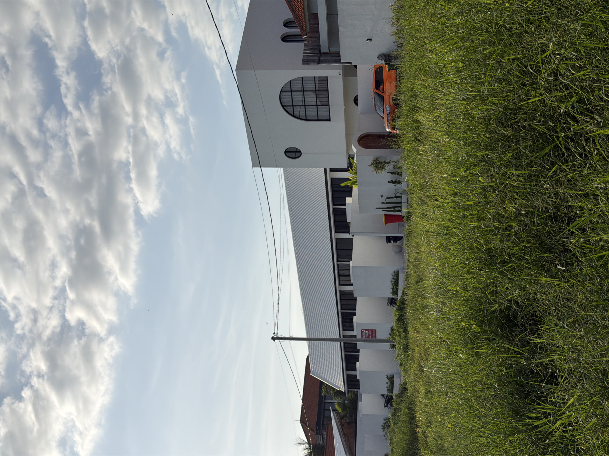The image shows a view of a white, modern building in North Kuta, Indonesia. The building features unique architectural elements, including arched windows and a metal roof. An orange vehicle is parked near the building. The building is adjacent to a field of green grass. The sky is partly cloudy. Overhead power lines are visible.