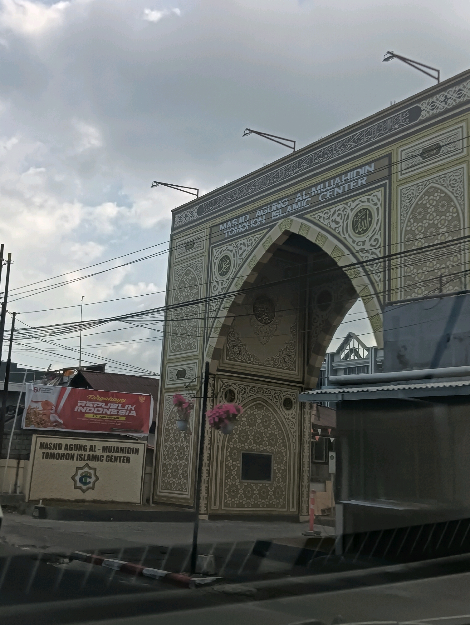 The image shows the archway of the Masjid Agung Al-Mujahidin Tomohon Islamic Center in Tomohon, Indonesia. The archway is adorned with intricate Islamic calligraphy and geometric patterns in shades of beige and white. Above the arch, text reads "MASJID AGUNG AL-MUJAHIDIN TOMOHON ISLAMIC CENTER." Two red and white striped bollards are visible in the foreground on a street. To the left of the archway is a banner that reads "Dirgahayu REPUBLIK INDONESIA 17 AGUSTUS." Several electrical wires crisscross the sky. The sky is overcast with gray clouds.