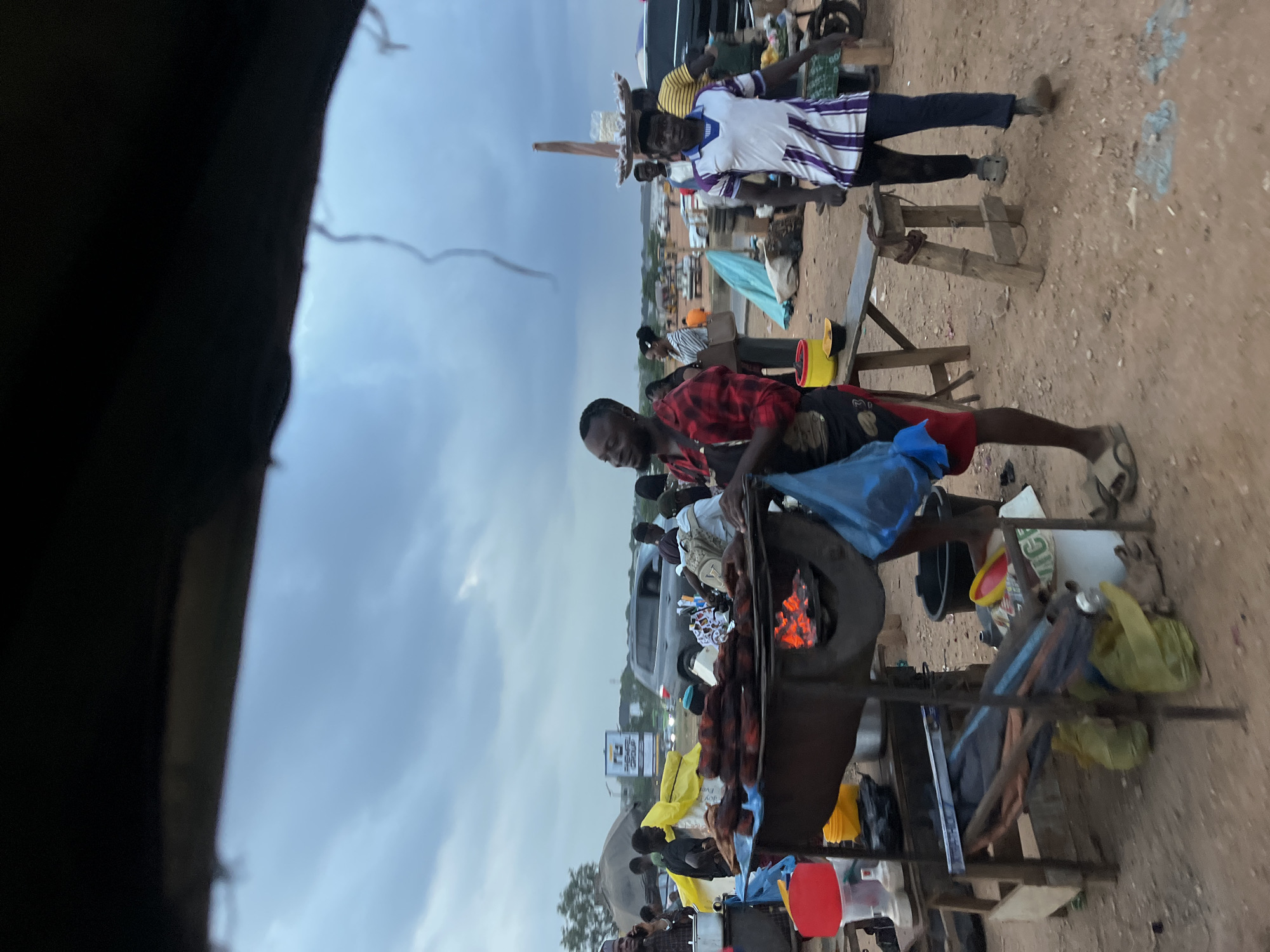 In Galadimawa Village, Nigeria, the image captures an outdoor scene near a dirt road. A man in a red plaid shirt stands at a makeshift grill, tending to food cooking over an open flame. Behind him, other people are visible. A man wearing a white shirt with purple stripes balances a round object on his head, and there are tables and chairs nearby. The environment appears to be a bustling outdoor market with various items scattered around. The sky is overcast with clouds. A car and several signs can be seen in the background, adding to the sense of activity in the area.