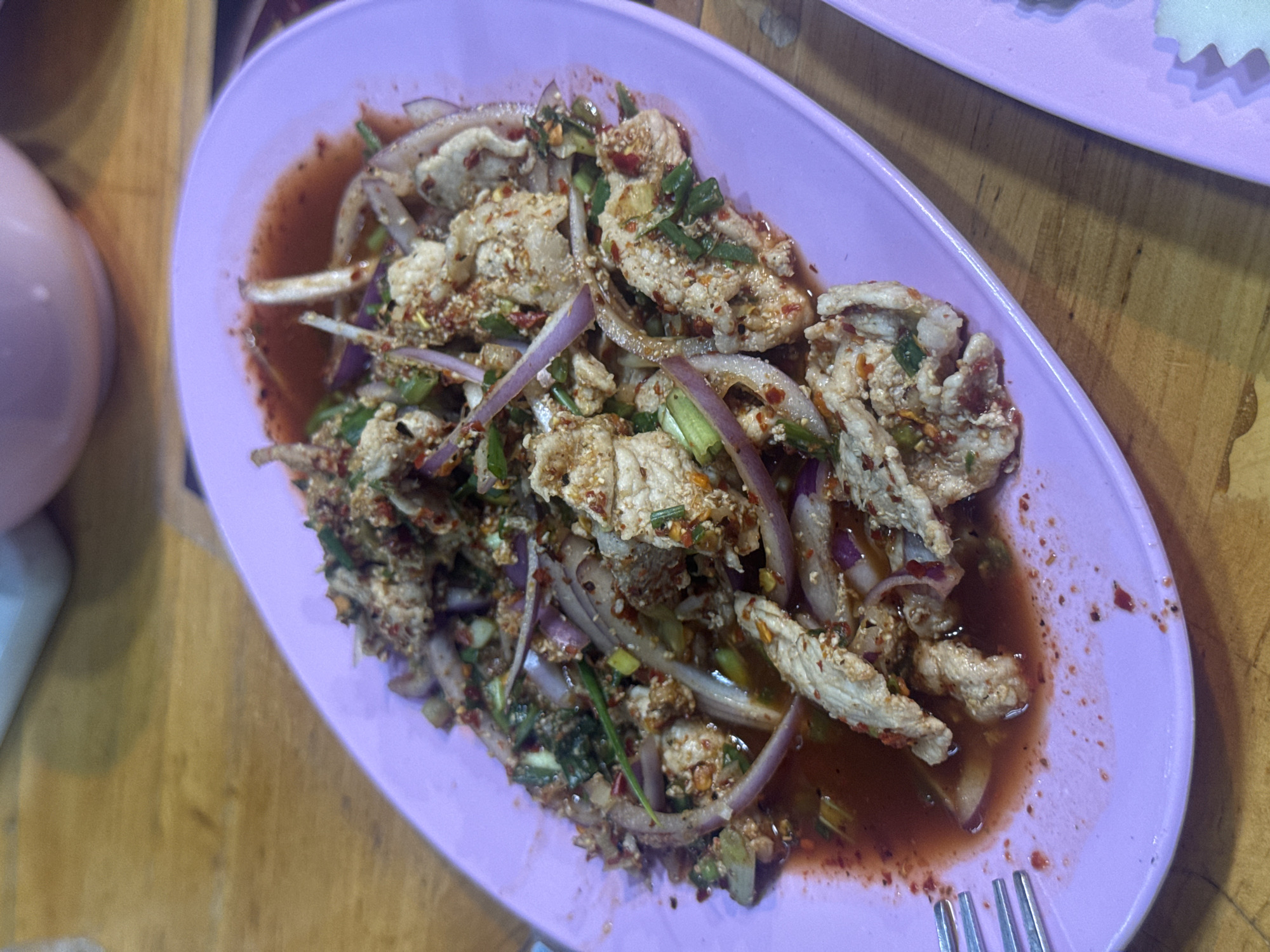 A plate of Larb Moo, a spicy Thai minced pork salad, sits on a wooden table in Pattaya City, Thailand. The dish consists of cooked minced pork, red onions, green onions, and herbs dressed in a spicy sauce. The plate is light purple and oval-shaped. A fork is visible on the lower right corner of the frame. Another plate is partially visible in the upper right corner. A light purple container is on the left side of the image.