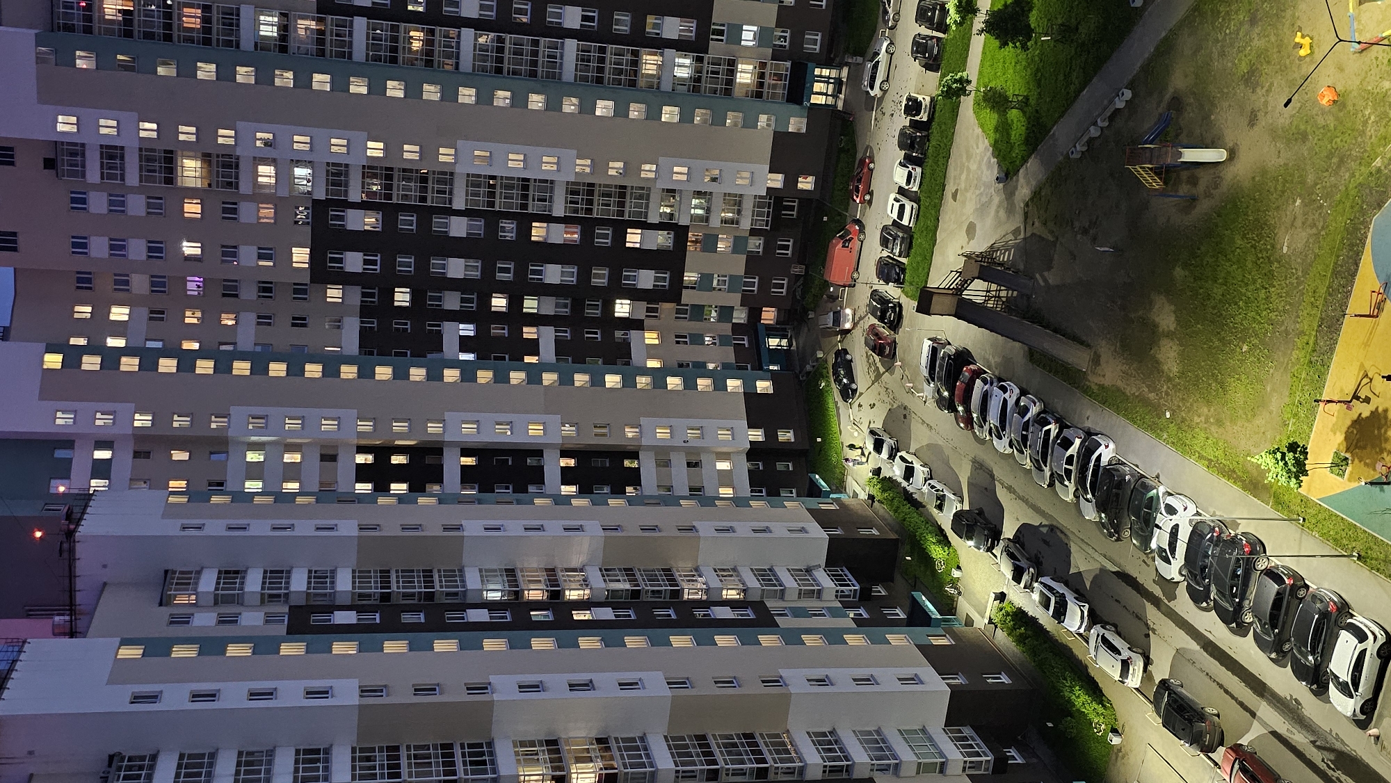 This is a high-angle, full-frame aerial view of a residential complex in Yekaterinburg, Russia at night. The scene is dominated by a large, multi-story apartment building with many illuminated windows, suggesting occupancy. In the foreground, a courtyard area is visible with a significant number of parked cars arranged in rows along a road and in designated parking spots. A small playground with a slide and some exercise equipment is also present. The lighting suggests it is evening or night.