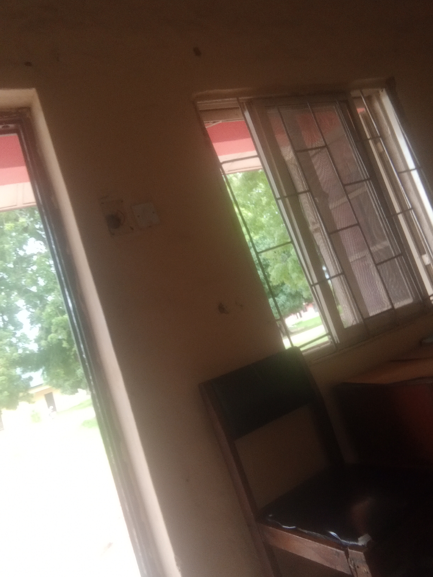 The image shows an interior view of a room with two windows. The left window is partially visible, revealing trees and a building outside. The right window has a metal grate and sheer curtains, with greenery visible beyond. A chair with a dark seat and back is placed in the foreground, partially obscuring a wooden surface. The walls appear to be painted a light color, and there are some electrical outlets visible on the wall between the windows. The lighting suggests it might be daytime, with natural light coming in from the windows. The overall scene suggests a simple room, possibly in a residential or institutional setting, in Jalingo, Nigeria. There is no discernible activity or event taking place, and no text is visible.