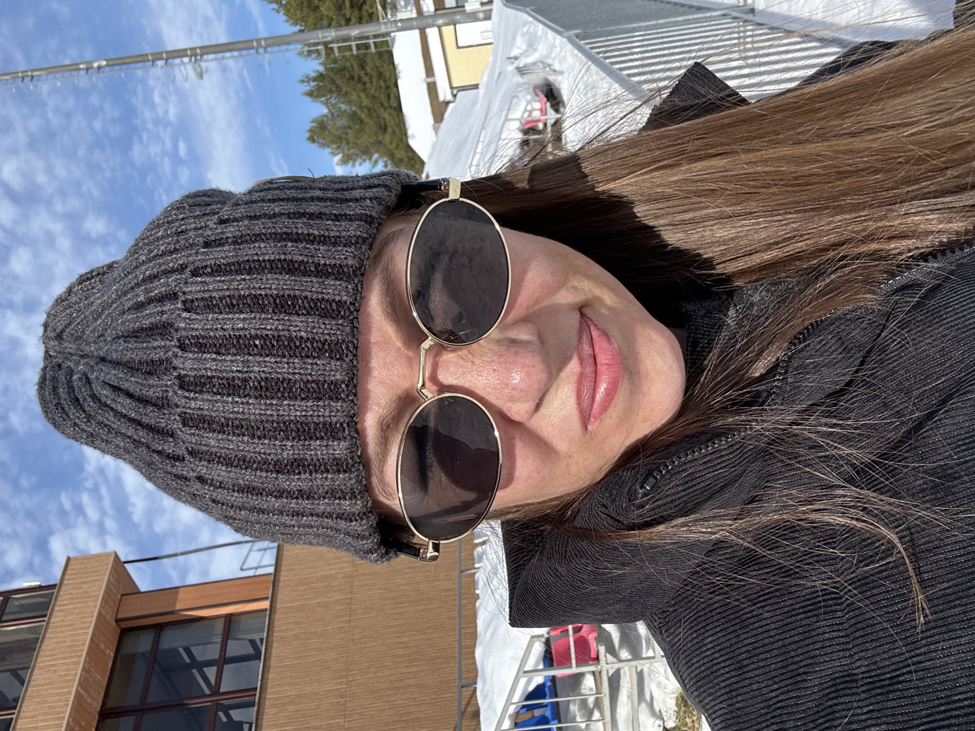 A female subject is depicted in a close-up, outdoor portrait taken on a clear, sunny day in Прикамский, Russia. She wears a dark grey ribbed knit beanie, gold-framed oval sunglasses with dark lenses, and a black ribbed corduroy jacket. Her long brown hair is visible along her right side. Her lips are slightly pursed. The background features a snow-covered environment. To the subject's left, a multi-story building with a light brown facade and large dark-framed windows is partially visible. To her right, a snowy landscape with pine trees and a tall metal mast or tower with ladder rungs is present. A white metal railing separates the foreground from the snow, behind which glimpses of colorful objects (red, blue) and another light-colored building are discernible.