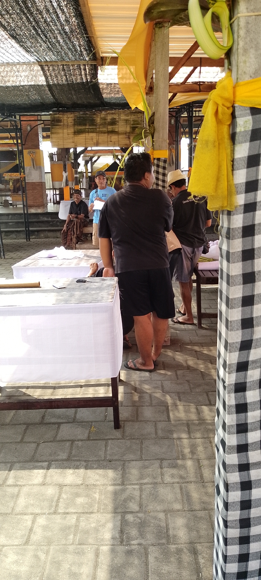 The image depicts an outdoor scene in Pekutatan, Indonesia, likely a gathering or event. The foreground is dominated by two rectangular tables covered with white cloths, one of which has a roll of fabric, scissors, and a phone on its surface. The tables are set on a paved ground made of square gray bricks, with shadows of sunlight indicating it is daytime.
In the midground, several people are present. On the left, a person in a dark shirt and sarong is seated, holding what appears to be a cup. To their right, a man in a blue t-shirt and a cap stands, holding a white paper. Further right, another man in a dark shirt and a hat is partially visible, facing away from the camera. Behind them, there's a building structure with a thatched roof and some bamboo blinds.
The background features more of the open-air structure, with wooden beams and a yellow fabric draped over a pillar. A large black and white checkered fabric is prominently featured on the right side of the image, obscuring part of the scene. The environment appears to be a semi-public space, possibly a temple courtyard or an event venue, characterized by the architectural style and decorative elements like the yellow fabric and the checkered cloth. The overall atmosphere suggests a casual gathering or preparation for an event.