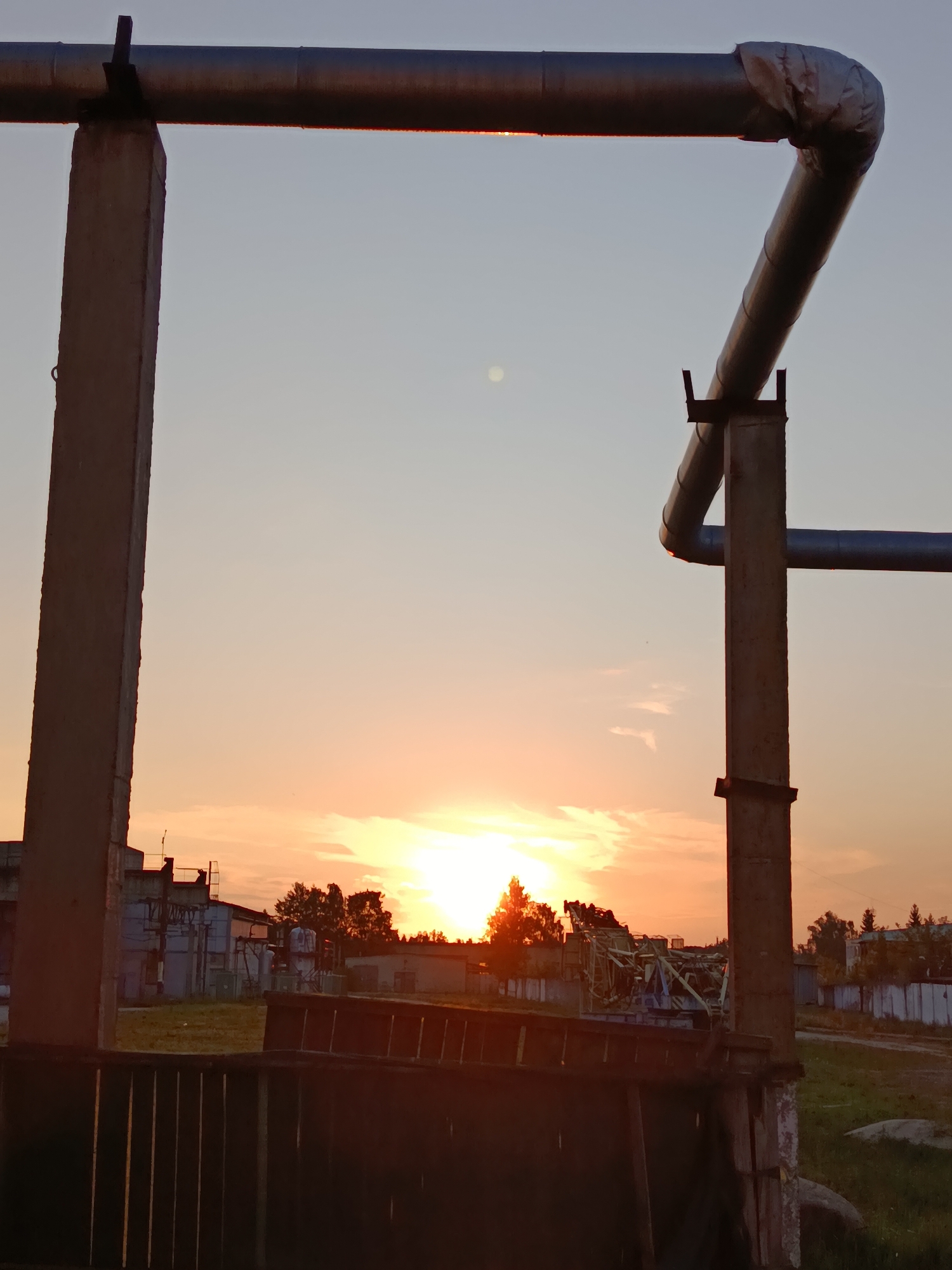 The image captures a sunset over an industrial area, possibly part of the city of Svietlahorsk, Belarus. Two large, grey concrete pillars support thick, insulated pipes that run horizontally and diagonally across the frame. The pipes are industrial in nature, with a visible join on the right side where one pipe bends sharply downwards. The sky is a gradient of soft blues and oranges, with the sun setting behind a line of trees in the distance.
Below the pipes and pillars, there is a low-lying industrial structure with buildings and what appears to be machinery or equipment. A dark, slatted fence or barrier is prominent in the foreground. The ground is covered in patchy grass, with some scattered rocks visible. The overall scene suggests an industrial complex at dusk, with the natural beauty of the sunset contrasting with the man-made structures. There are no discernible individuals present in the image.