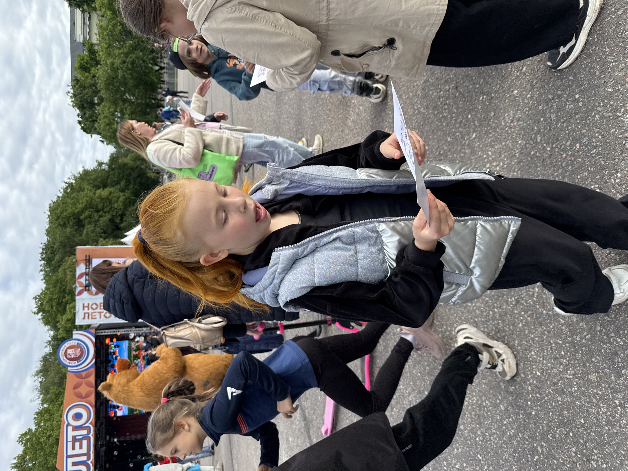 A slightly overhead shot shows a group of people on a gray asphalt surface, likely a street or large paved area. The composition is dominated by a young red-haired girl in the foreground who is holding a piece of paper with text or drawings, appearing to read or study it. She is wearing a dark top, black pants, and a silver puffer vest.
Several other people are partially visible behind her. One is wearing a beige jacket. Further back, a banner for a "New Summer" festival is visible, with a large teddy bear figure in front of a stage where an event appears to be taking place. There are bubble machines next to the stage.
The presence of the "New Summer" banner, written in Russian, suggests the location is in Russia. Given that the photograph was associated with the city of Veliky Novgorod, Russia, it can be inferred the event and setting are located in this city.