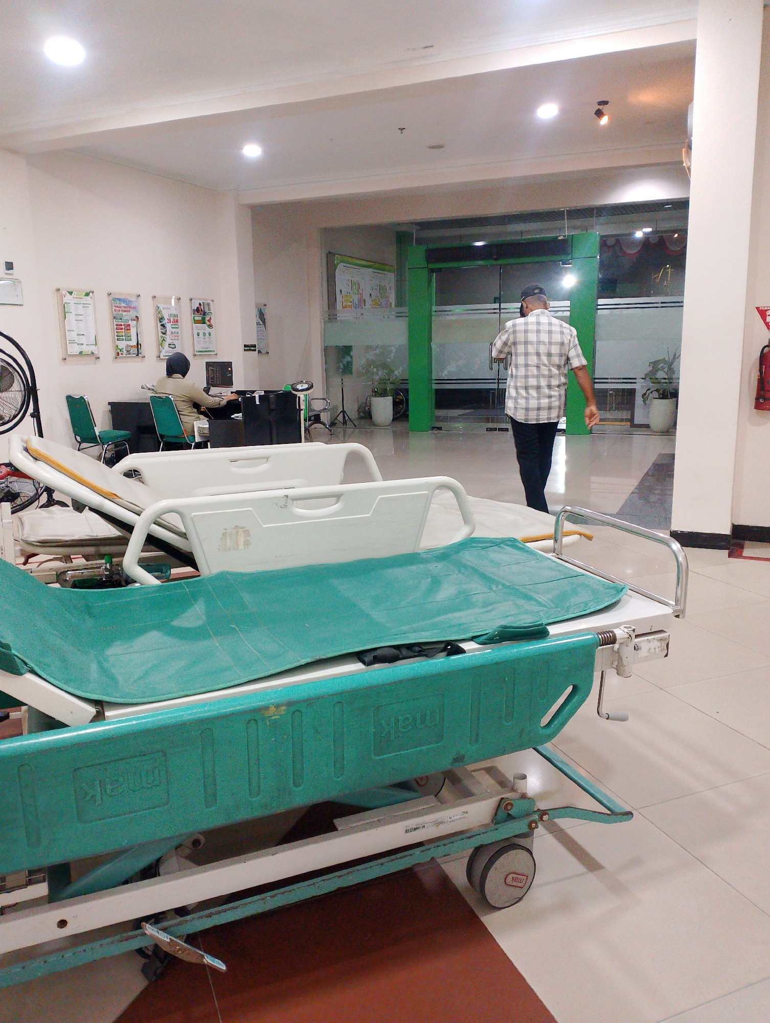 The image depicts a hallway in a medical facility, possibly a hospital or clinic, in Sidoarjo, Indonesia. In the foreground, two hospital beds are visible, with one prominently displaying a green mattress and a white plastic frame. The beds are on wheels, suggesting they are movable. The hallway features tiled flooring and bright overhead lighting. On the left side, a black desk with a computer and chairs is occupied by a person wearing a headscarf, seemingly working. Further down the hallway, a man in a checkered shirt and dark pants is walking away from the camera, heading towards a set of glass doors. The doors lead to an area with green accents and visible plants, indicating a reception or waiting area. The environment appears clean and organized, typical of a healthcare setting. A red fire extinguisher is mounted on a white column on the right side of the image.