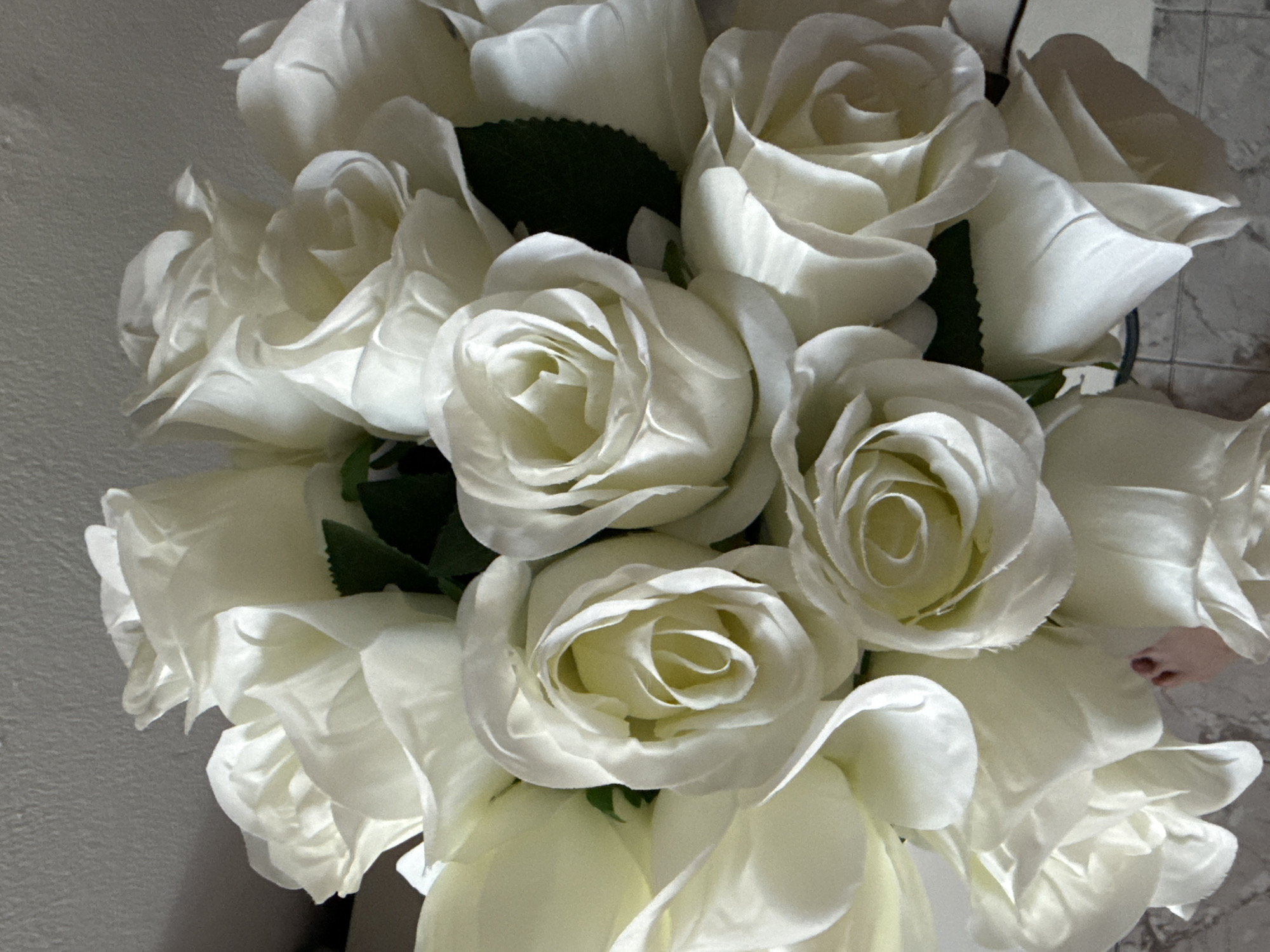 A close-up, top-down shot captures a bouquet of artificial white roses. The roses exhibit detailed petal formations, with some buds tightly closed and others open, revealing their layered interiors. A few dark green artificial leaves are interspersed amongst the blooms. The background features a textured light-colored wall, casting subtle shadows. The lower portion of the image shows a blurred surface with a tiled pattern, possibly a countertop or floor. A small, bare foot is partially visible in the lower left quadrant, stepping slightly into the frame, with toes pointing upwards. There is no indication of Bat Yam, Israel in the image.