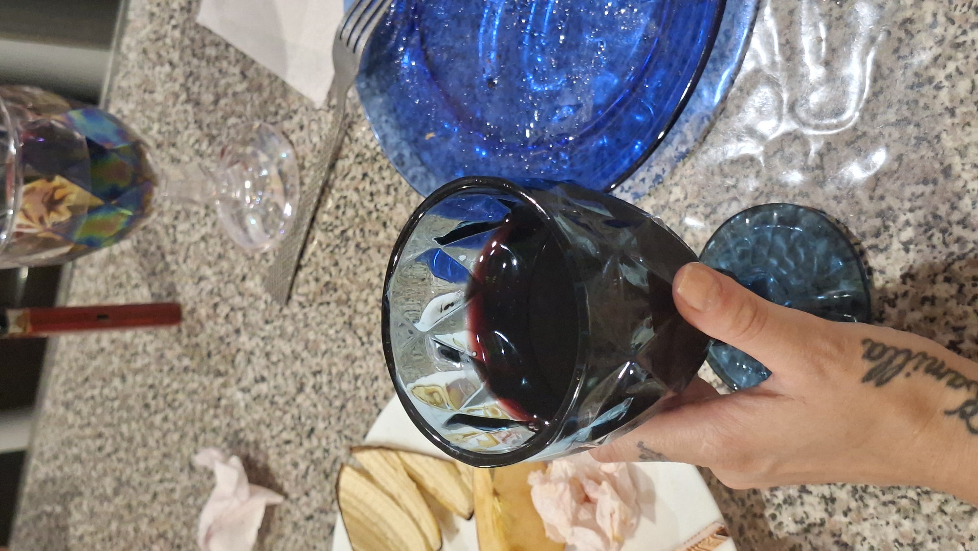 A close-up, eye-level shot shows a person's left hand holding a dark blue, faceted wine glass filled with red wine. The glass is positioned in the foreground, with the person's thumb resting on the stem and fingers wrapped around the bowl. A tattoo of the name "Camilla" is visible on the back of the hand.
In the background, on a speckled countertop, are several objects. To the left, a white plate holds a portion of food, appearing to be dried banana chips and some shredded cheese. A crumpled light pink napkin is also on the plate. Further back, a red object, possibly a vape pen, stands upright. To the right of the wine glass is a shiny blue plate with remnants of food. A silver fork is also visible on the countertop. Another clear wine glass with iridescent hues filled with red wine is in the upper right background. The overall environment suggests a casual meal or gathering. The location is Myadzyel, Belarus.
