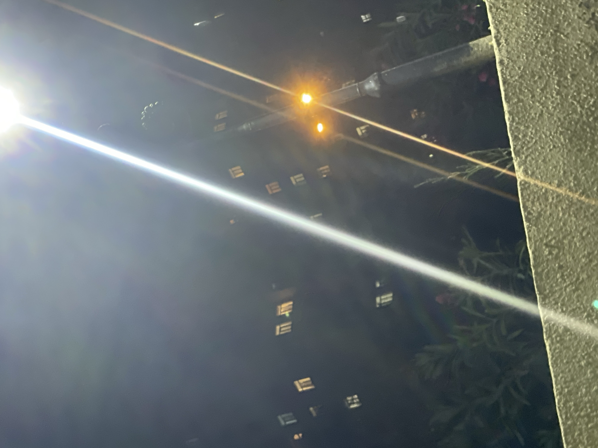 This is a nighttime photograph taken in the city of Sumgayit, Azerbaijan, depicting a dimly lit street with a prominent lamppost in the foreground. The lamppost has two lights, one brighter orange-yellow and a smaller one below it. Streaks of light from the lamppost cut diagonally across the image. In the background, a dark building with numerous lit windows is visible. Several trees and foliage are present in the lower portion of the image, with a textured, light-colored wall or ledge in the immediate foreground. The overall atmosphere is dark, with artificial lighting creating lens flare and light scattering effects.