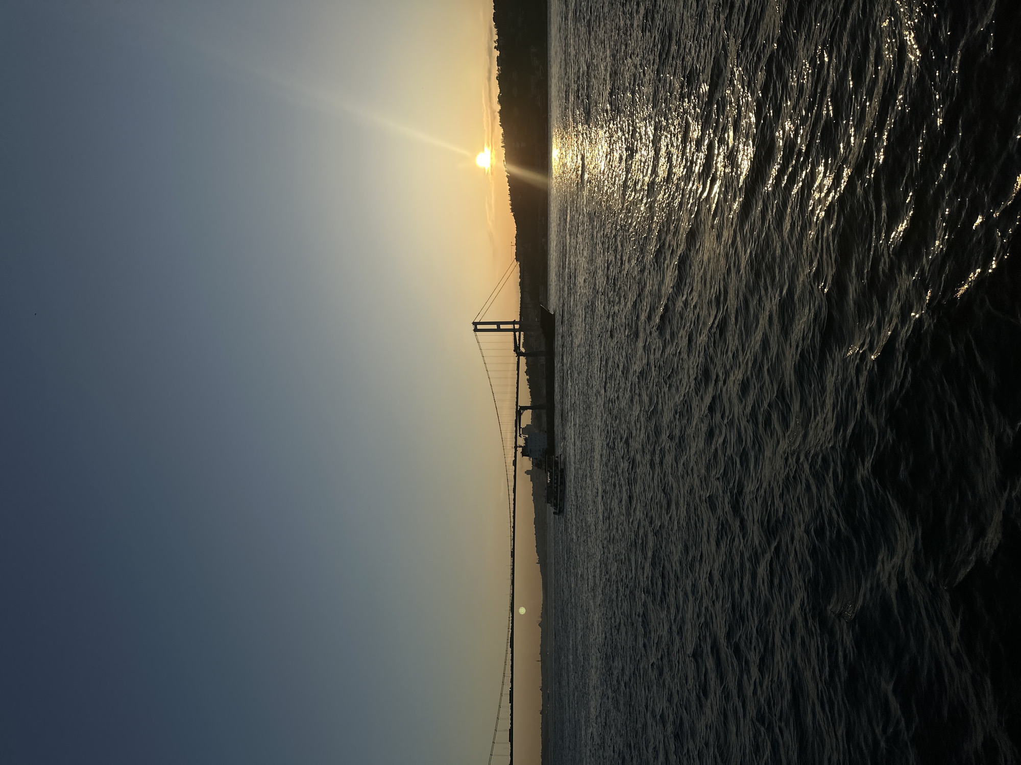 The image depicts a sunset over the Bosphorus Strait in Üsküdar, Turkey. The sun is setting behind a tree-lined landmass. A suspension bridge spans the strait. The water surface reflects the sunlight, creating a shimmering effect.