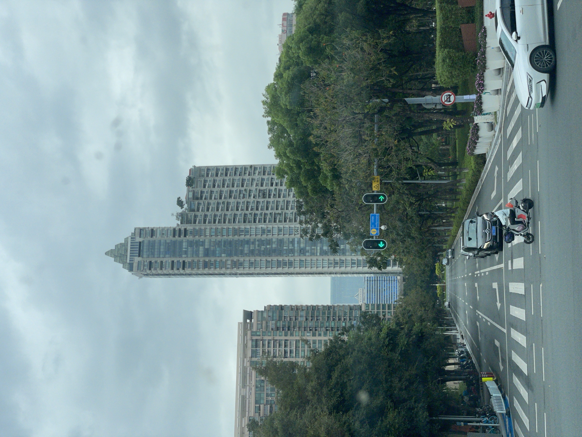 The image depicts a street scene in Yuexiu District, China. Tall buildings are visible on the left, partially obscured by trees. A multi-lane road runs through the scene, with a crosswalk and directional arrows painted on the asphalt. Two cars, one white and one dark, are present on the road. A motorcycle-like vehicle is also on the road. A street sign with a prohibition symbol is visible, along with green traffic lights. The sky is overcast, suggesting a cloudy day. Trees and greenery line the side of the road, and bicycles are parked alongside the road.