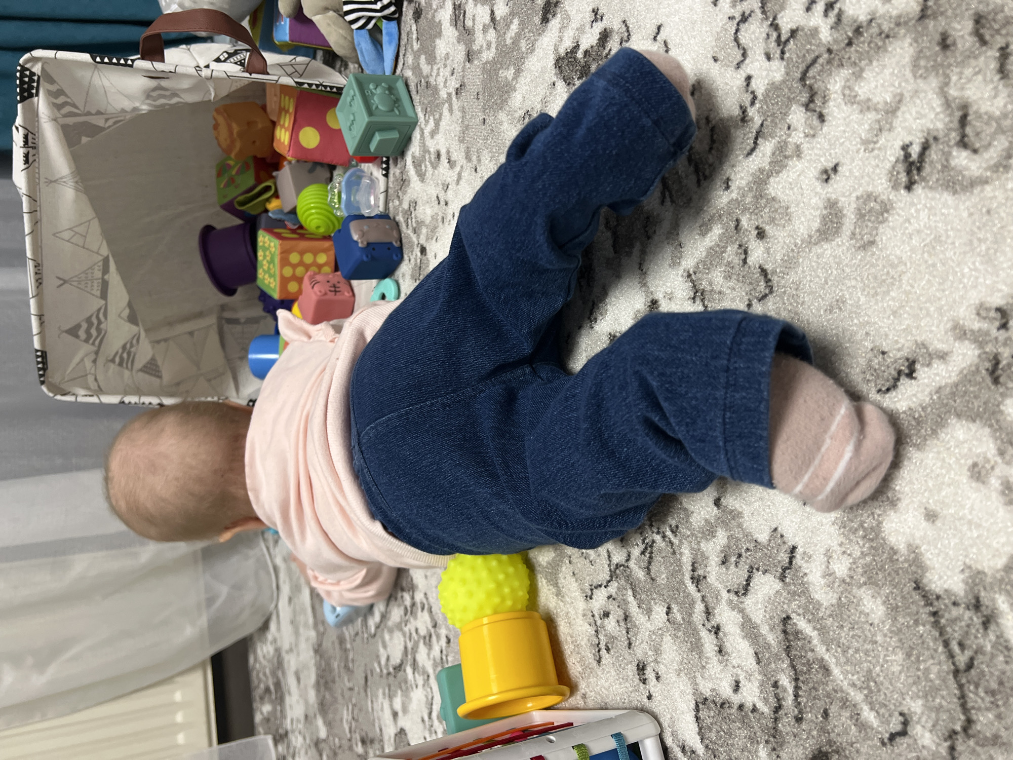 A baby lies on a gray patterned rug, wearing blue jeans and pink socks. The baby is positioned near a toy basket filled with colorful blocks and other toys. A yellow and green toy is located near the baby's torso. The photo was likely taken indoors. There is no information present that indicates the image was taken in Chaikovsky, Russia.