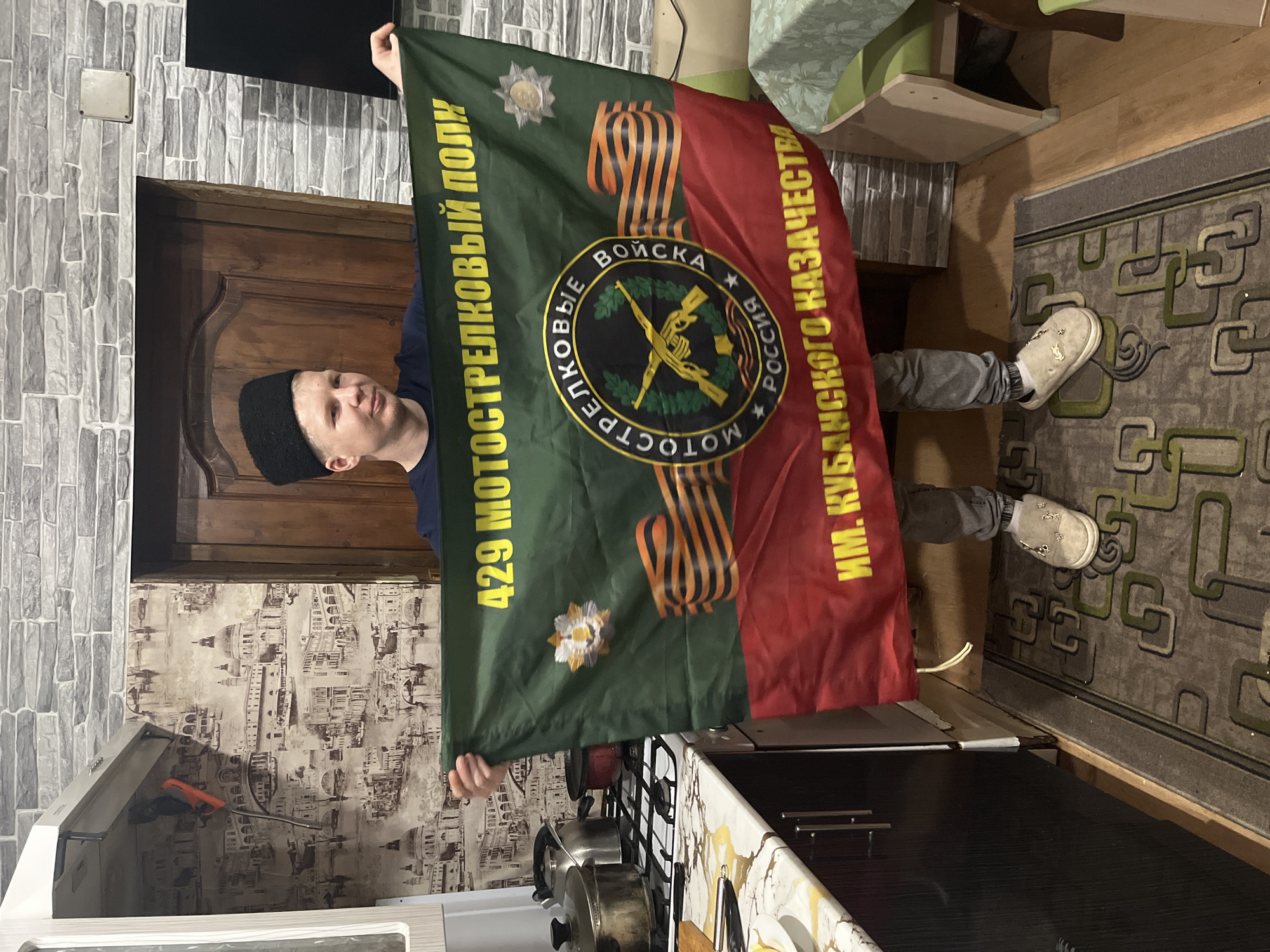 A person is holding a flag indoors. The flag is green and red with a central emblem and text in Russian, indicating it is the flag of the 429th Motorized Rifle Regiment named after the Kuban Cossack Army. The person is wearing a black hat and grey pants. The setting appears to be a kitchen. There is a gas stove, wallpaper featuring city scenes, and a patterned rug on the floor. There is a door visible behind the person and the wall appears to be made of brick. It is not possible to ascertain from the image that this is Vesyoloye, Russia.