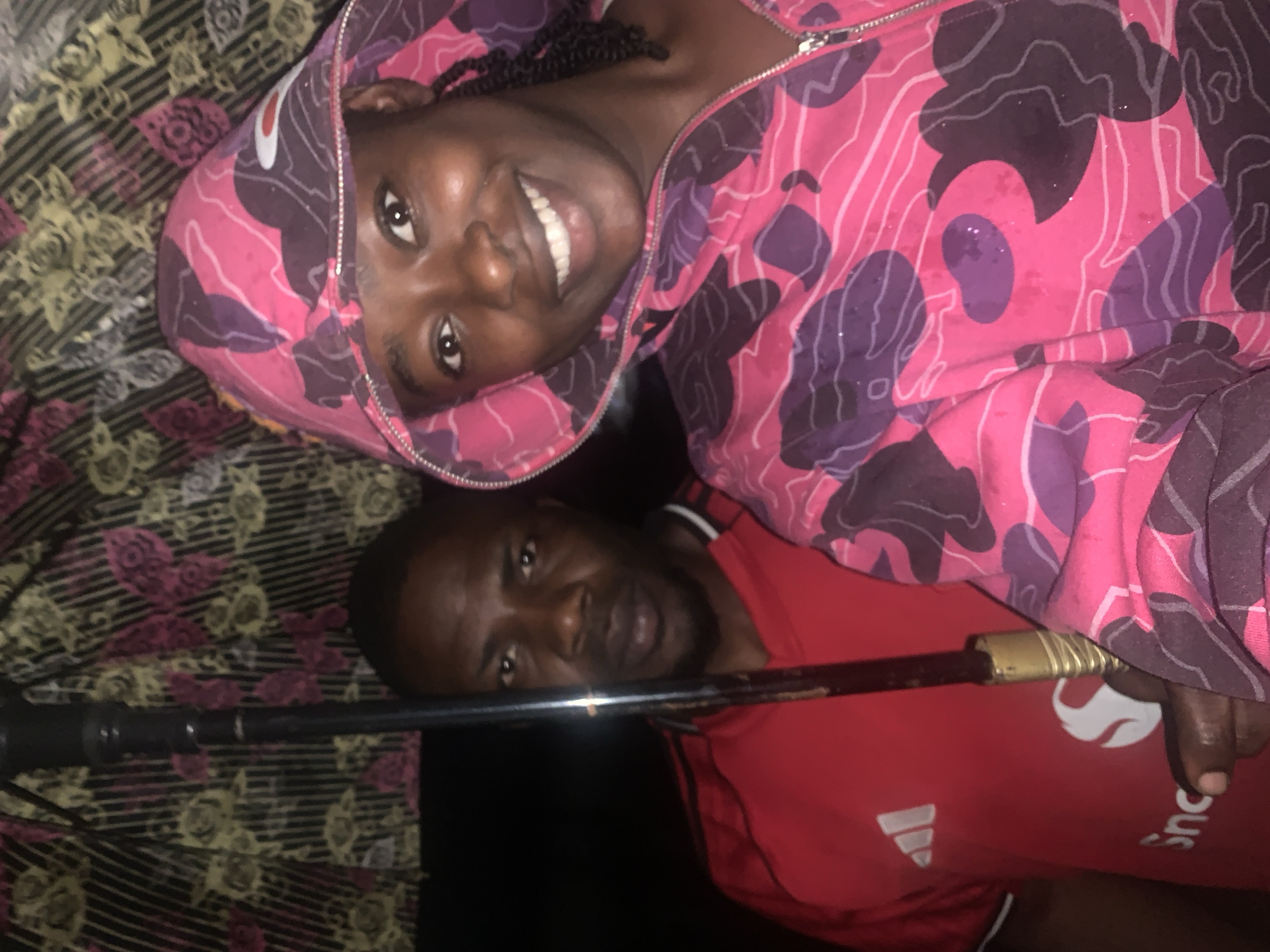 This image, captured in Rafin Par, Nigeria, features two individuals posing in what appears to be an indoor setting. In the foreground, a woman lies on her back, looking directly at the camera with a broad smile, conveying happiness. She is wearing a distinctive pink and purple camouflage-patterned hooded jacket, which is zipped up. Her black braided hair is visible around her face.
Behind and slightly below her, a man is also present, looking towards the camera with a neutral expression. He is wearing a red short-sleeved shirt, which clearly displays a white Adidas logo (three stripes) on the front, and partially visible white text that begins with "Sinc...".
A dark, metallic pole-like object runs horizontally across the mid-ground, partially obscuring the man's face and extending towards a gold-colored end near his hand. The background consists of a patterned fabric, possibly a blanket or bedspread, featuring black stripes overlaid with floral or geometric designs in shades of yellow, green, and pink/purple. The lighting suggests an indoor setting, likely evening or nighttime, with direct illumination on the subjects. Both individuals are in a relaxed, casual posture.