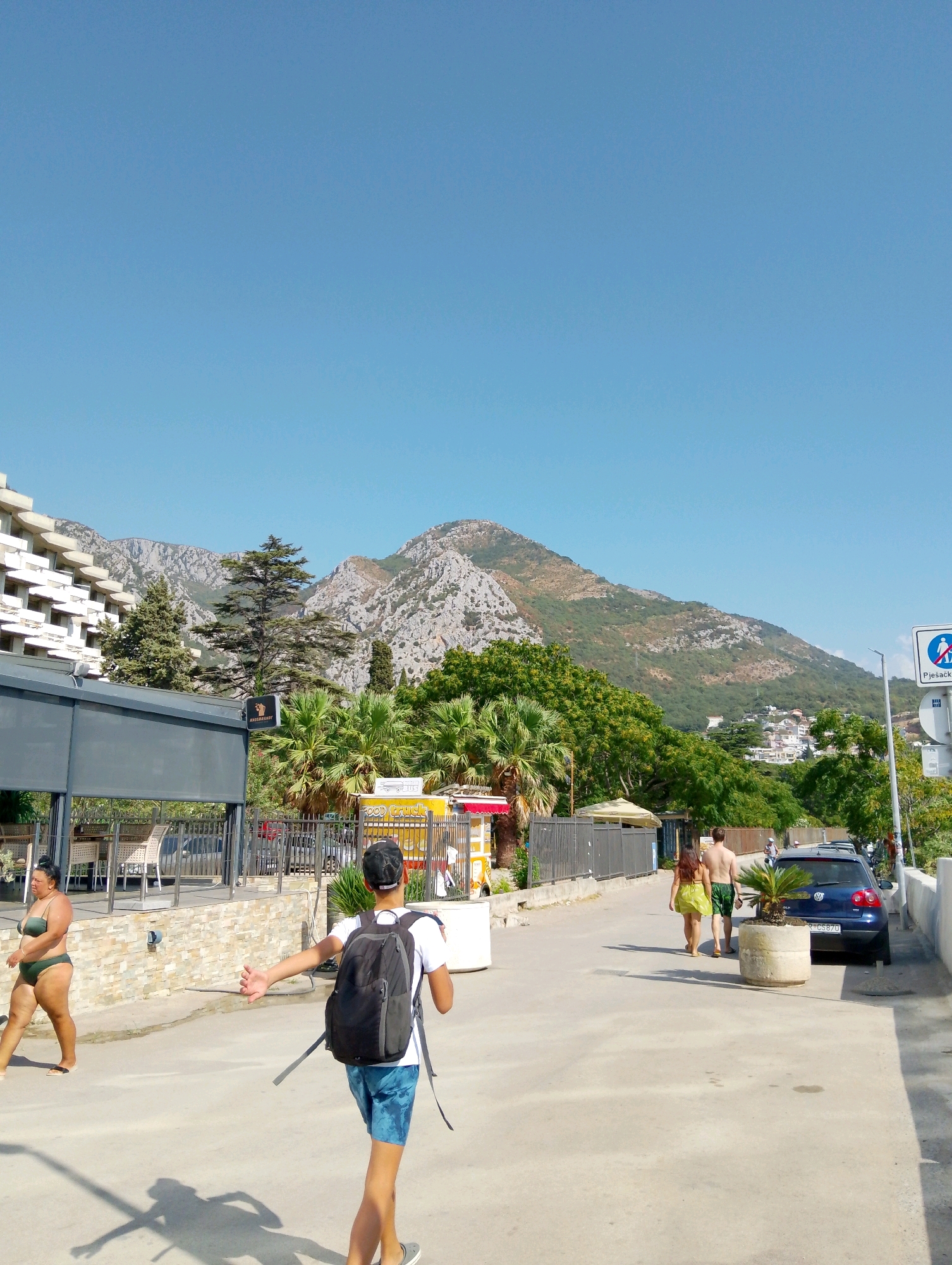 In Sutomore, Montenegro, under a clear blue sky, a paved road runs parallel to the Adriatic Sea. On the left, a multi-story building with balconies overlooks the scene, with a shaded outdoor seating area beneath it. A woman in a green bikini walks away from the camera. In the center, a young man wearing a white t-shirt, blue patterned shorts, and a black backpack is walking away from the camera, with his right arm extended outwards as if gesturing or reaching. To the right, a couple walks away from the camera, the woman in a yellow dress and the man shirtless. A blue car is parked on the right side of the road. Lush green trees and palm trees are visible throughout the scene, with a large, rocky mountain range forming the backdrop. The overall atmosphere is bright and sunny, indicative of a summer day.