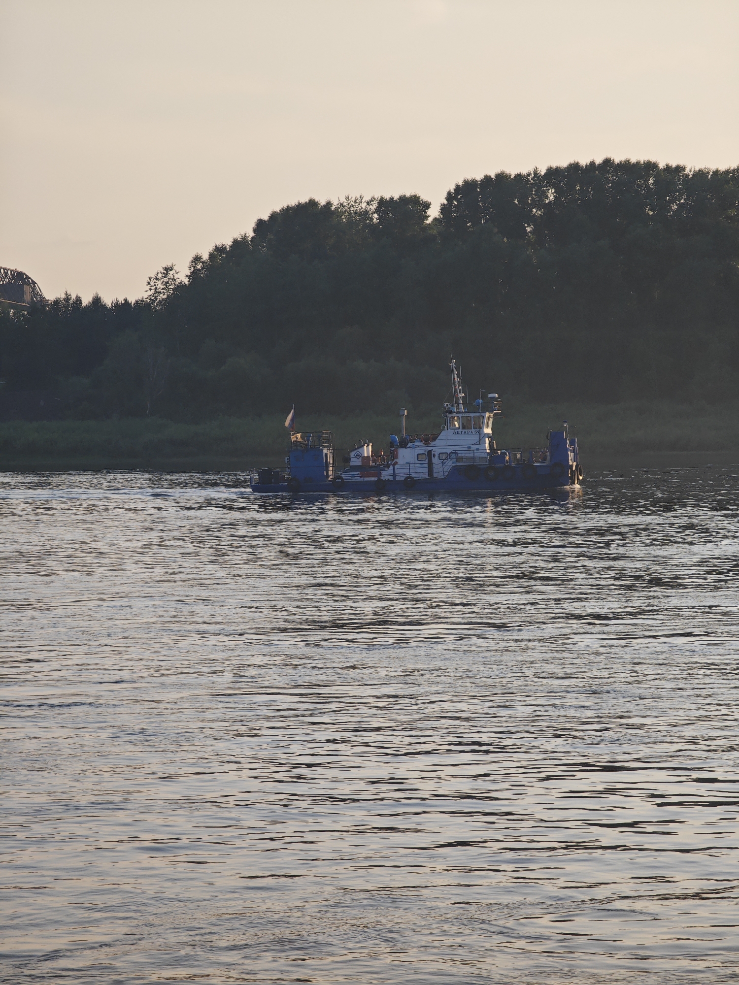 A blue and white ferry boat with a Russian flag sails on a wide, calm river at dusk. The boat has a cabin structure with windows and railings. The river is surrounded by a dense forest on the far bank, and a faint outline of a bridge can be seen on the left side of the image, suggesting an urban or semi-urban setting within or near Lesosibirsk, Russia. The sky is a pale, overcast hue, indicative of twilight. The water's surface reflects the soft light, creating ripples and streaks of shadow. There are no visible people on the deck of the boat.