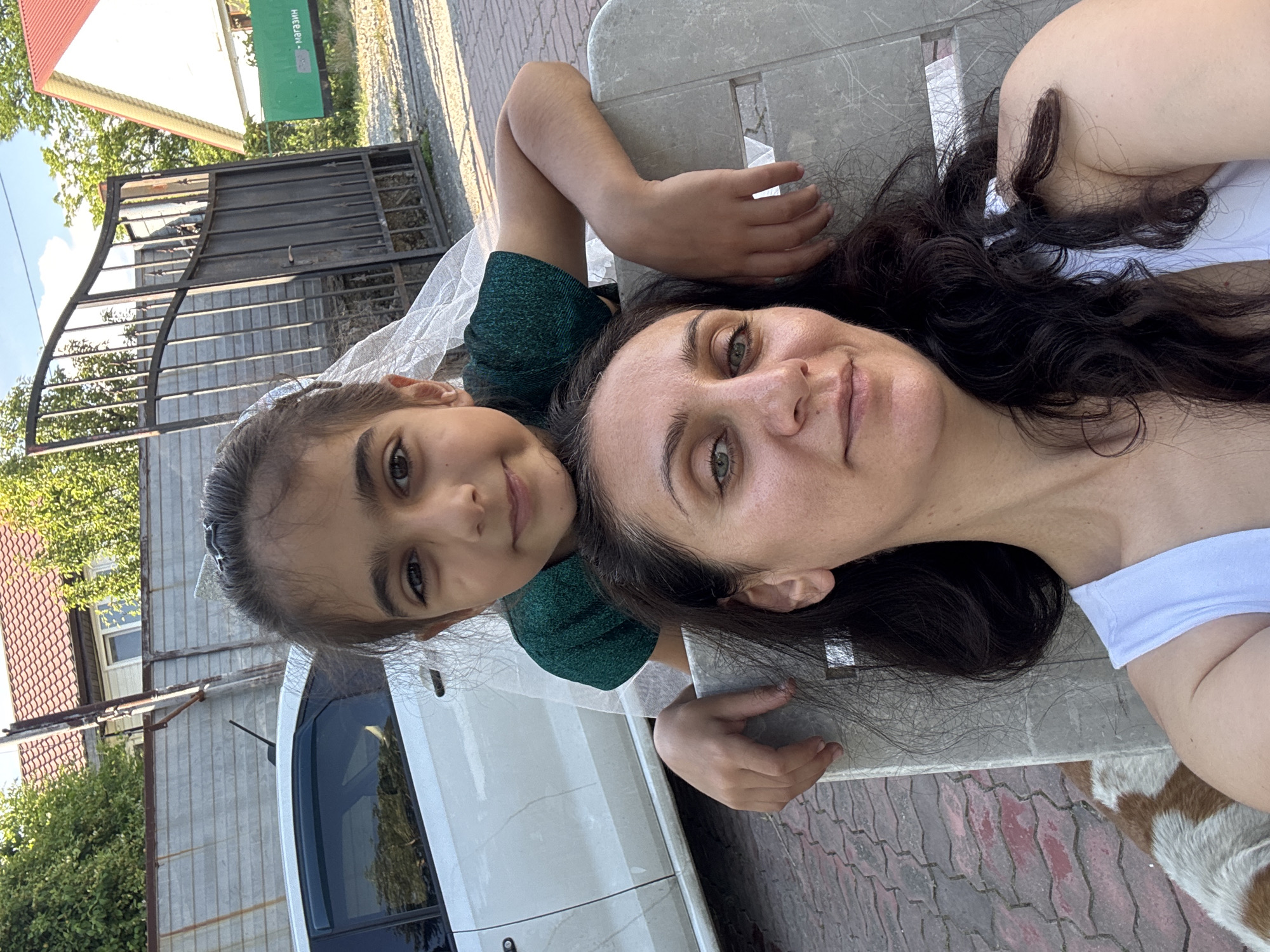 In this vertical, close-up selfie, a woman and a young girl lie down together on a light gray picnic table outdoors. The woman, with dark, wavy hair and green eyes, is on the right side of the frame, wearing a white top. The girl, with dark hair and brown eyes, is on the left, leaning on her arm. She wears a dark green sparkly dress and a white veil or hair accessory. Their faces are close together, and they both appear to be smiling or looking relaxed.
Behind them, a black metal gate and a fence are visible, with a white car parked nearby. Lush green trees and a building with a red roof are also in the background. The ground is paved with interlocking bricks. The lighting suggests it is daytime, likely sunny, as indicated by the clear sky and shadows.
There is text visible on a green sign in the upper left portion of the image, reading "МАГАЗИН" which translates to "SHOP" in Russian.
The image evokes a sense of warmth and connection between the two subjects. The setting appears to be a residential area or a park, given the presence of the house, car, and picnic table.