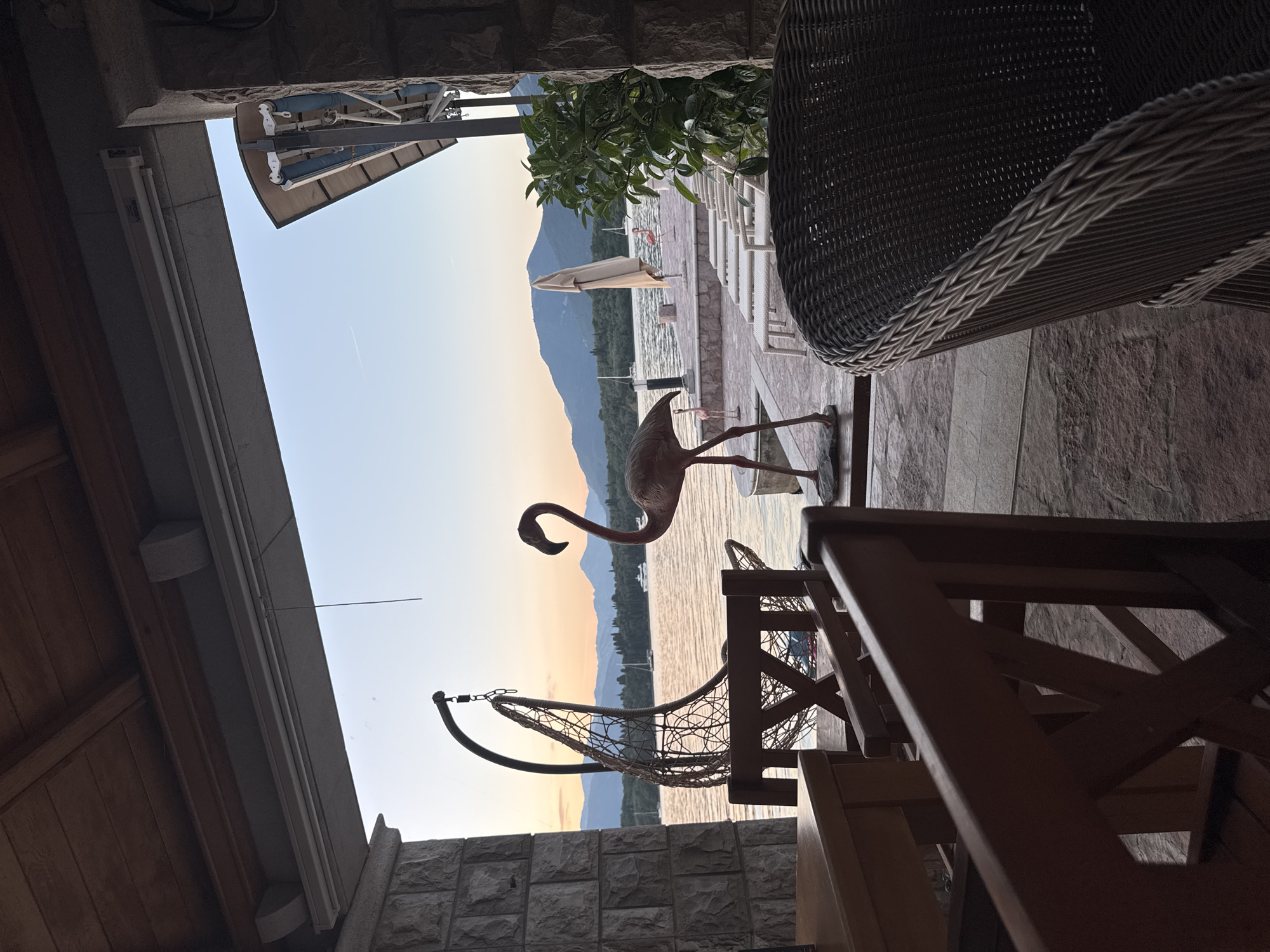 The scene captures a view from an outdoor covered space, likely a patio or veranda, in Bogišići, Montenegro. Through the opening, a body of water extends towards a mountainous backdrop under a sky transitioning from light to dusk. A decorative metal flamingo stands on the paved surface near the opening. In the foreground, partially obscuring the view, are elements of outdoor furniture, including wicker chairs, a wooden chair, and a hanging macrame chair. The structure housing the patio appears to be made of stone and wood. In the distance, white lounge chairs and a partially opened sun umbrella are visible along the waterfront.