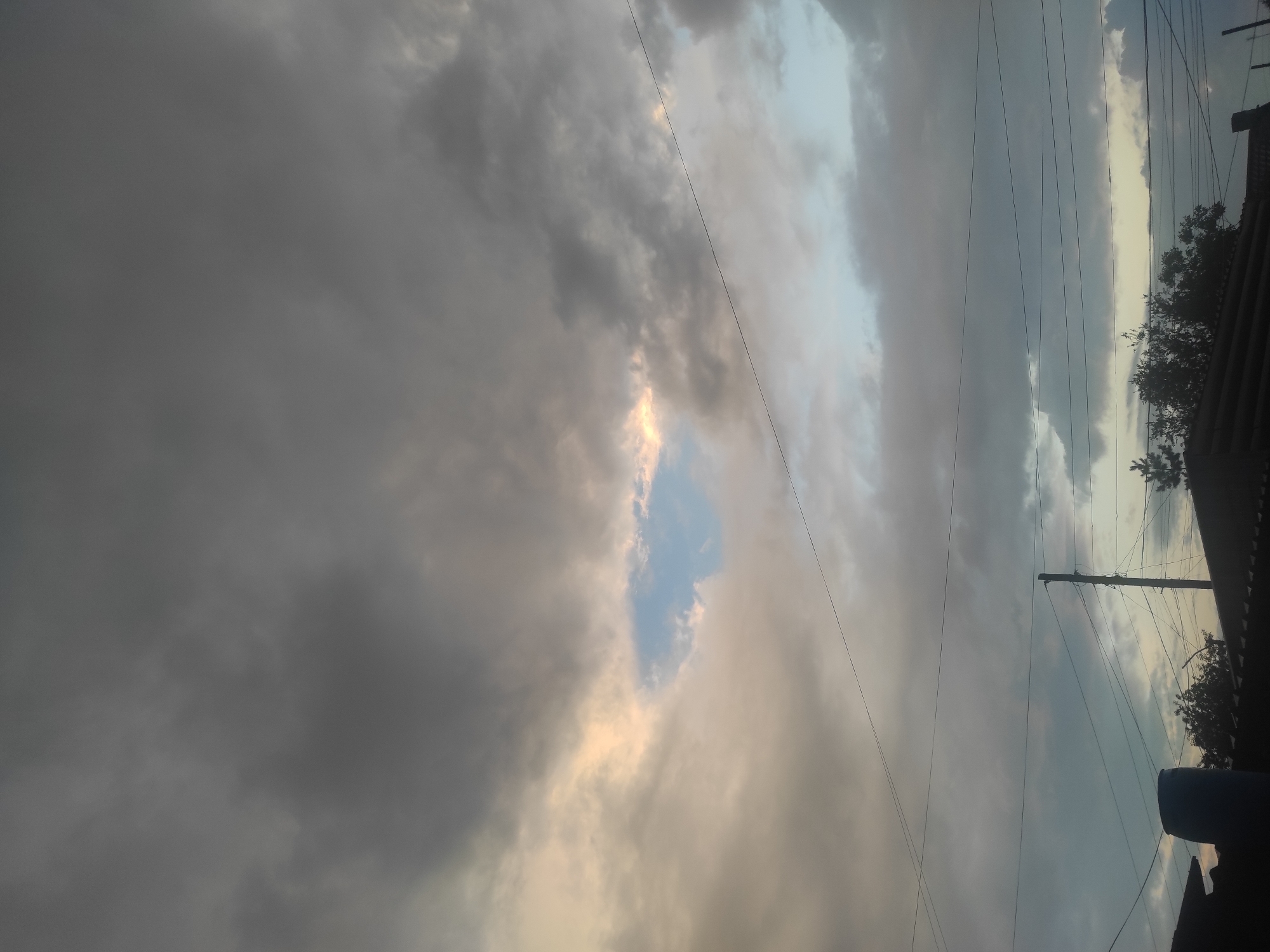 The image depicts an overcast sky with patches of sunlight breaking through the clouds, illuminating a small area of blue sky. Several utility wires crisscross the frame, supported by wooden poles. In the foreground, the rooftops of buildings are visible, along with the silhouette of a large blue barrel and trees. The scene suggests an outdoor setting in a residential or semi-urban area. The weather appears to be changing, with a mix of dark and light clouds. There are no people or animals visible in the image. The overall impression is one of a cloudy day with a hint of sunlight, typical of the city of Shakhtinsk, Kazakhstan.