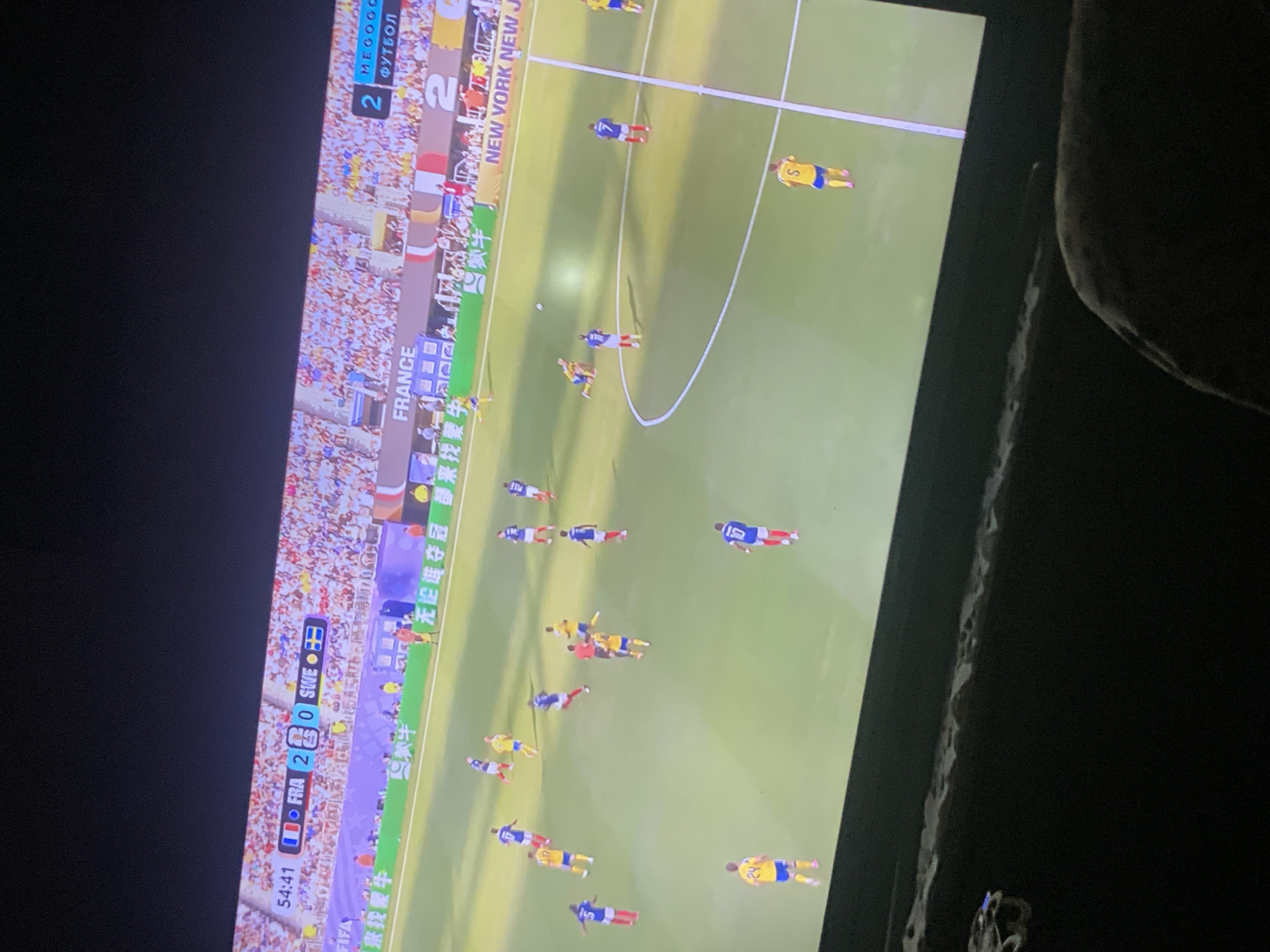 The image shows a soccer match displayed on a television screen. The score board indicates France is playing Sweden, with France leading 2-0. The time is 54:41. Players in blue and white uniforms (France) and yellow and blue uniforms (Sweden) are visible on the green field. The crowd in the stadium is blurred in the background. The television is positioned in a dark room, with a portion of the screen reflecting some ambient light. There is no indication of Zastavna, Ukraine in the image.