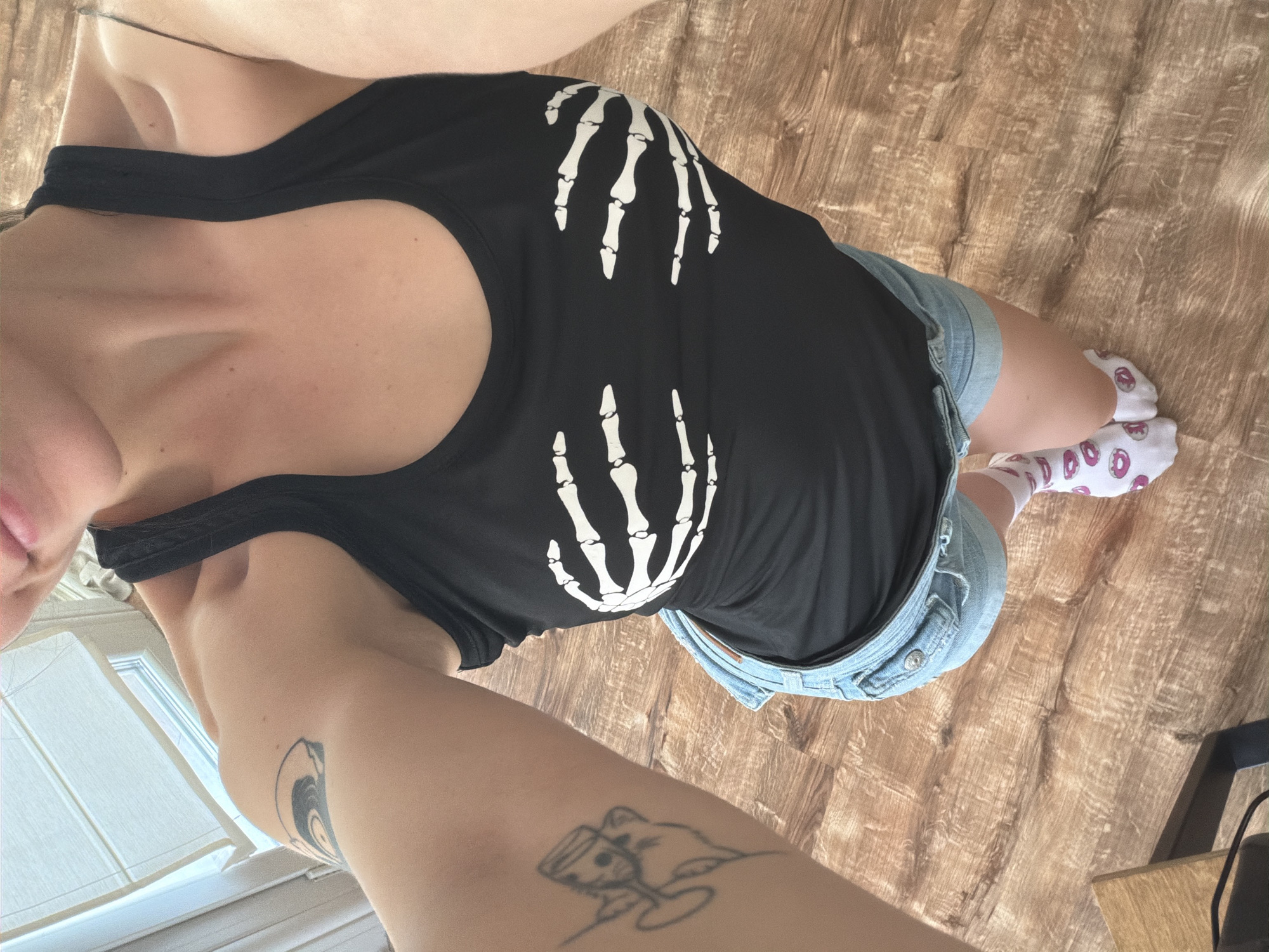 A woman is standing in a room with wooden flooring, taking a selfie from a high angle. She is wearing a black tank top with a skeleton hand print on the chest, light-wash denim shorts, and white socks with pink donut prints. Her left arm is visible on the left side of the image, showing two tattoos: one on the upper arm and another on the forearm. The background shows a window with blinds and a sliver of a wall.