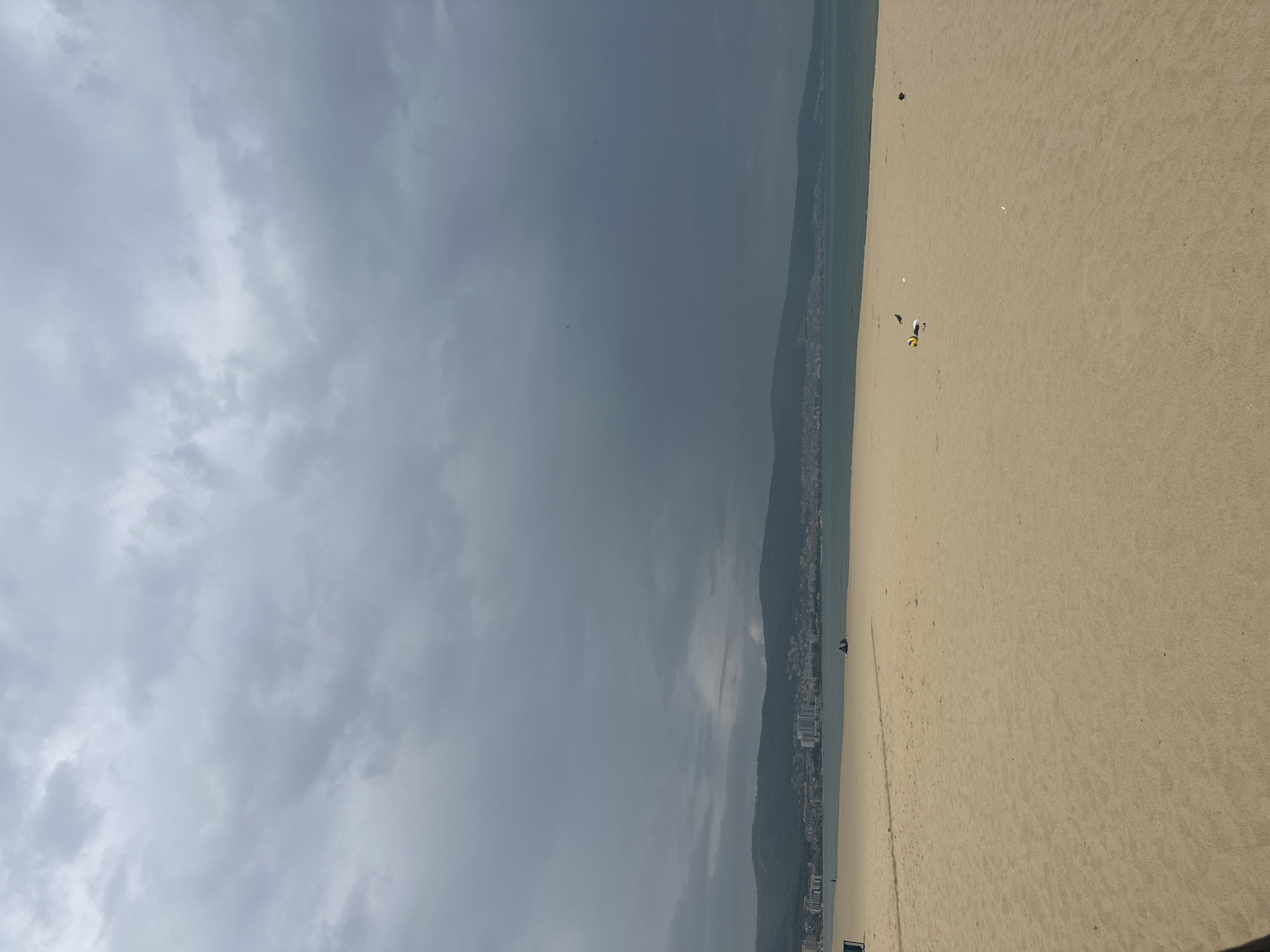 The image shows a beach in Nesebar, Bulgaria on a cloudy day. The beach is sandy and extends along the coast with a line of buildings and greenery on the side where land meets water. The sky is overcast with grey clouds. There are some people on the beach, sparsely distributed.