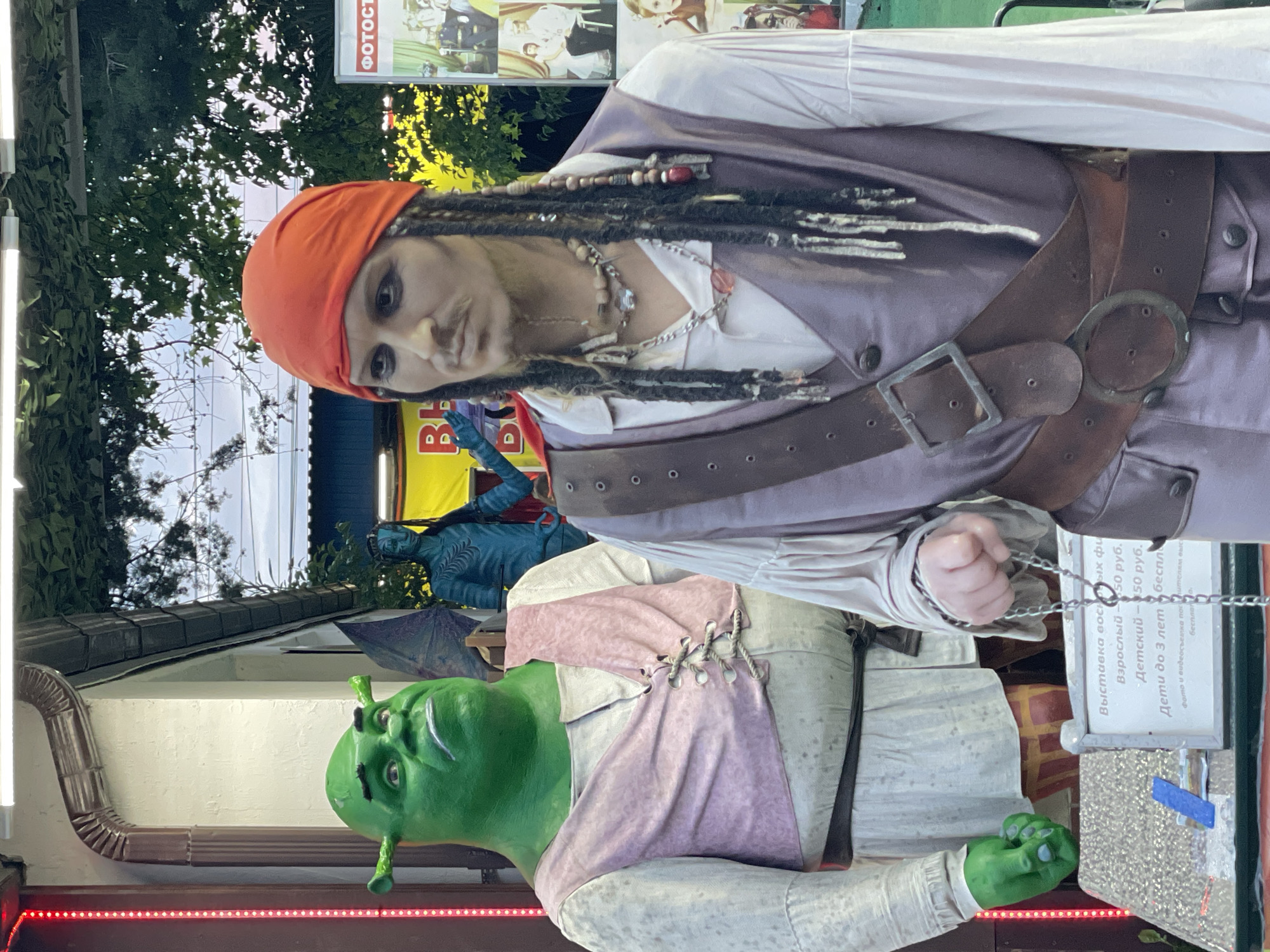 The image shows figures of Jack Sparrow, Shrek, and Avatar, photographed outside in the city of Sochi, Russia. Jack Sparrow, with a red bandana and dreadlocks, is positioned at the top. Shrek, with his signature green skin, stands below him. Behind them is a blue Avatar figure. A sign with Russian text and prices is visible, likely indicating fees for photos or exhibitions. The background includes trees, a building with lighting, and a photo booth sign.