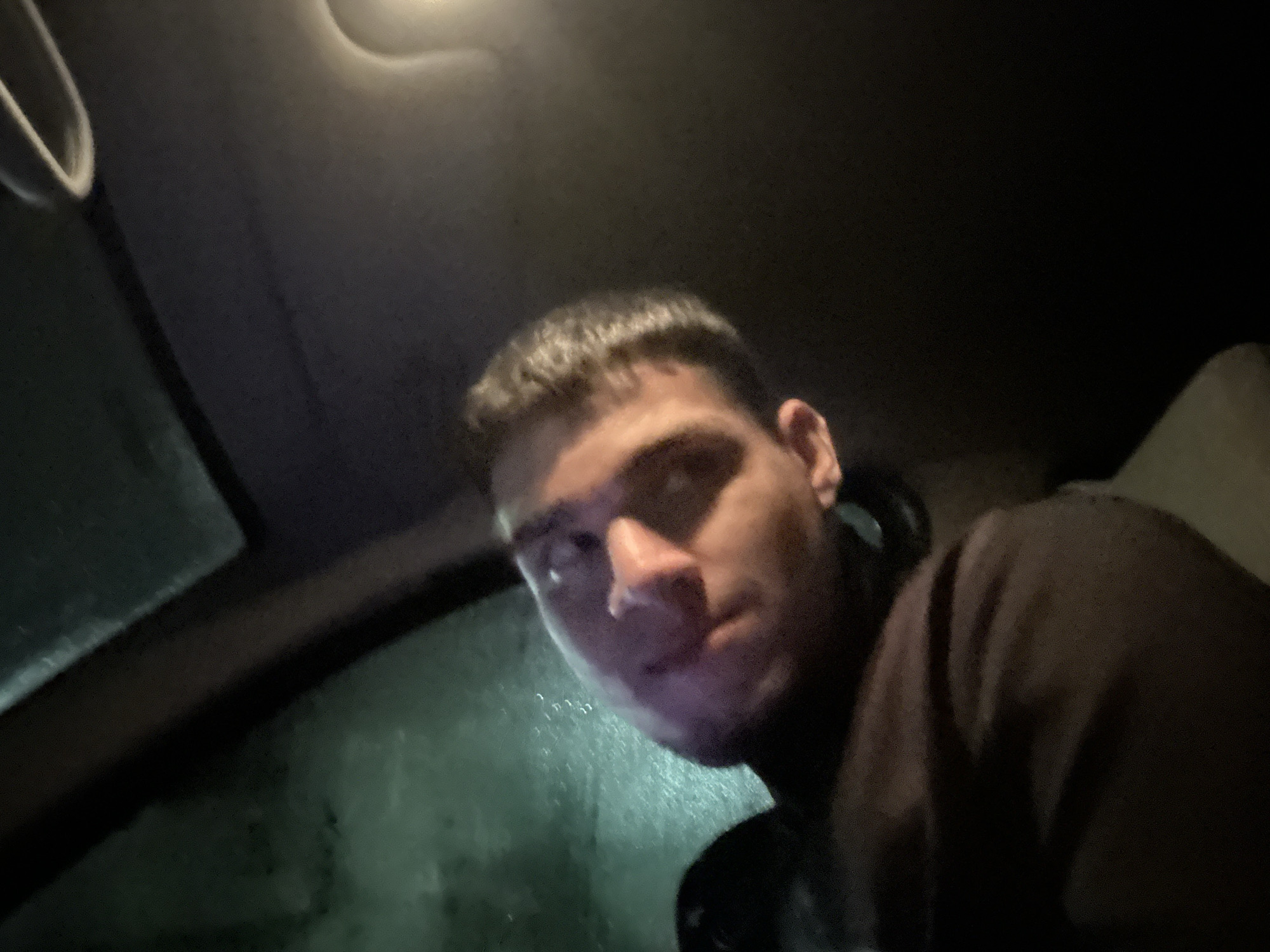 The image depicts the interior of a vehicle at night in Unknown, Unknown. A male individual, positioned in the lower-center-right, is partially visible from the chest up, facing the camera. He has short, dark hair and is wearing a dark-colored top. His gaze is directed towards the camera. The vehicle's dark headliner is visible at the top, with a light source or reflection in the upper-left quadrant. The lower-left portion of the frame shows a window, likely the windshield, with visible condensation or water droplets. The scene is dimly lit.